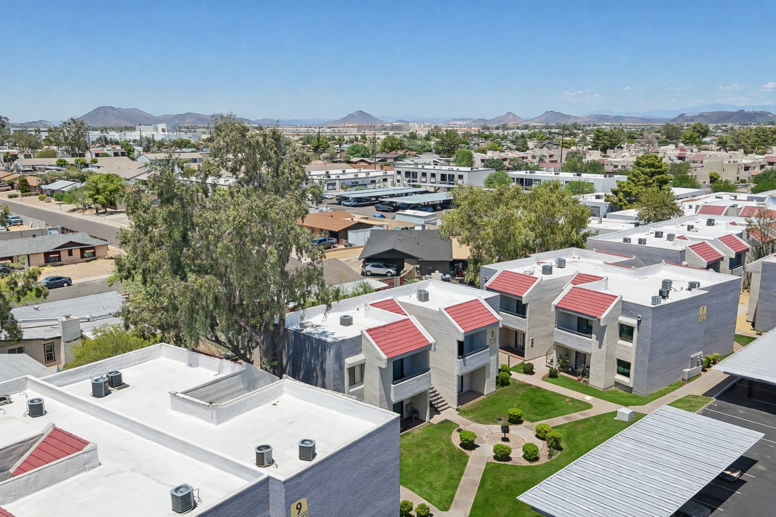 Aerial view of a residential area featuring flat-roofed apartment buildings with red accents. Surrounding greenery and a parking area are visible, along with a broader cityscape in the background, including distant mountains and various buildings across the horizon. The sky is clear and sunny.