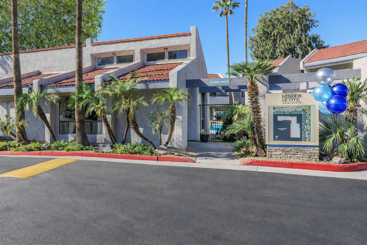 Entrance to a residential complex named "Hidden Cove," featuring a modern building design with palm trees and landscaping. A large sign displays the name of the complex. Balloons in blue and silver decorate the entrance area, and there's a swimming pool visible through the entrance.