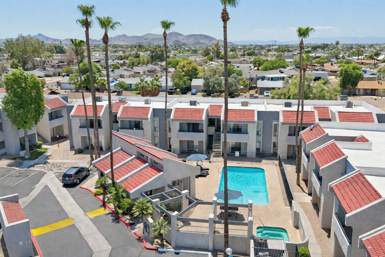Aerial view of a modern residential complex featuring multiple buildings with red-tiled roofs surrounding a swimming pool. Palm trees are visible along the edges, and scenic mountains are in the background. The area is filled with greenery and residential homes.