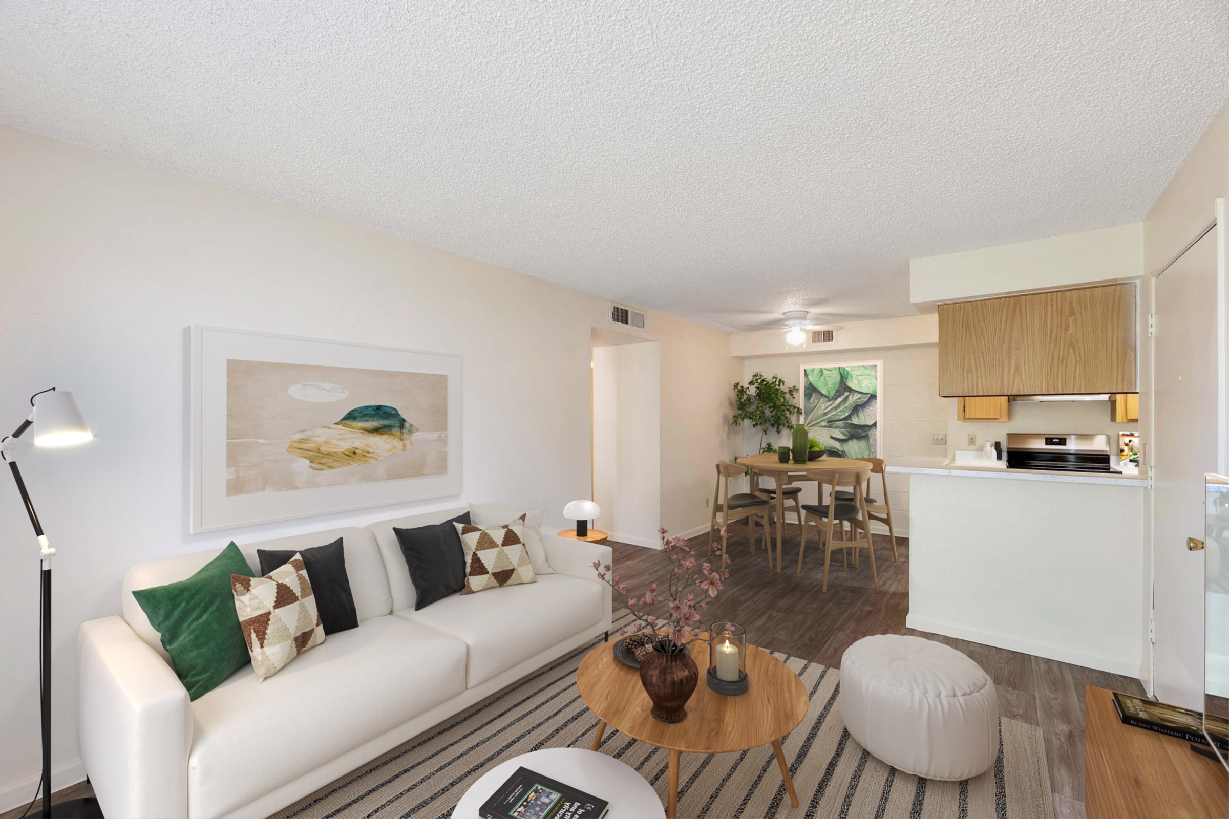 A bright, modern living room featuring a white sofa adorned with decorative pillows, a round coffee table with candles and a plant, and a cozy pouf. In the background, there's a dining area with a wooden table and chairs. A large window allows natural light, enhancing the welcoming atmosphere.