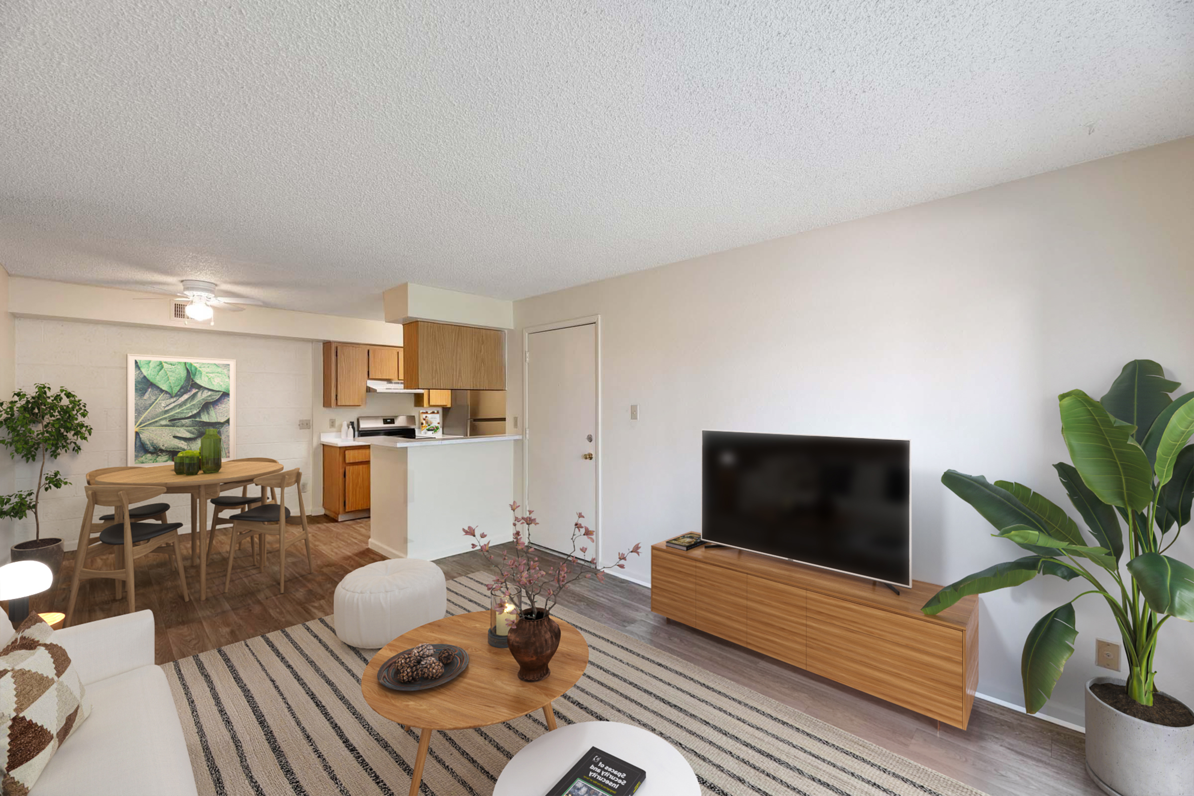 A modern living space featuring a cozy seating area with a coffee table, a large television on a wooden stand, and a dining area with a round table and chairs. The room has a light color scheme, plants for decoration, and a textured ceiling, creating a welcoming atmosphere.