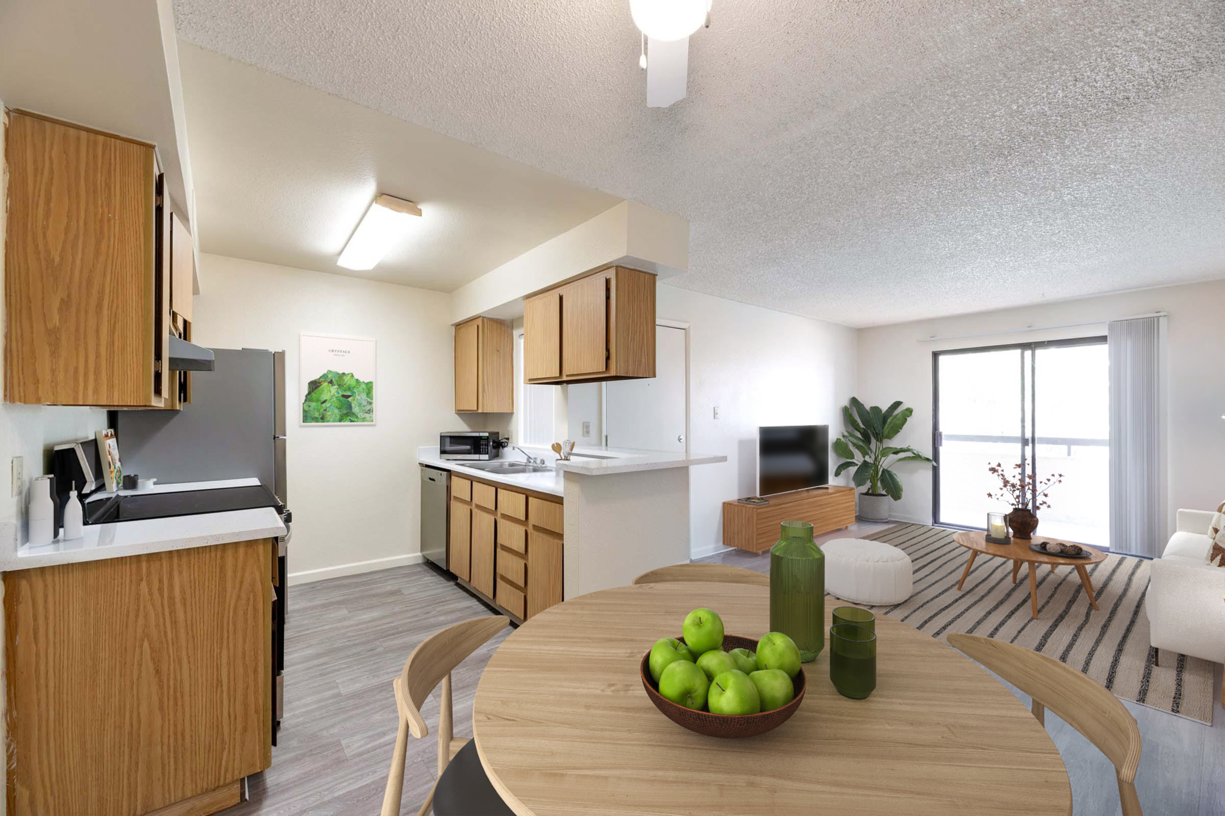 A bright and modern living space featuring an open kitchen with wooden cabinets, a dining table with green apples, and a comfortable sitting area with a light-colored sofa and plants. Large windows allow natural light to fill the room, creating a welcoming atmosphere.