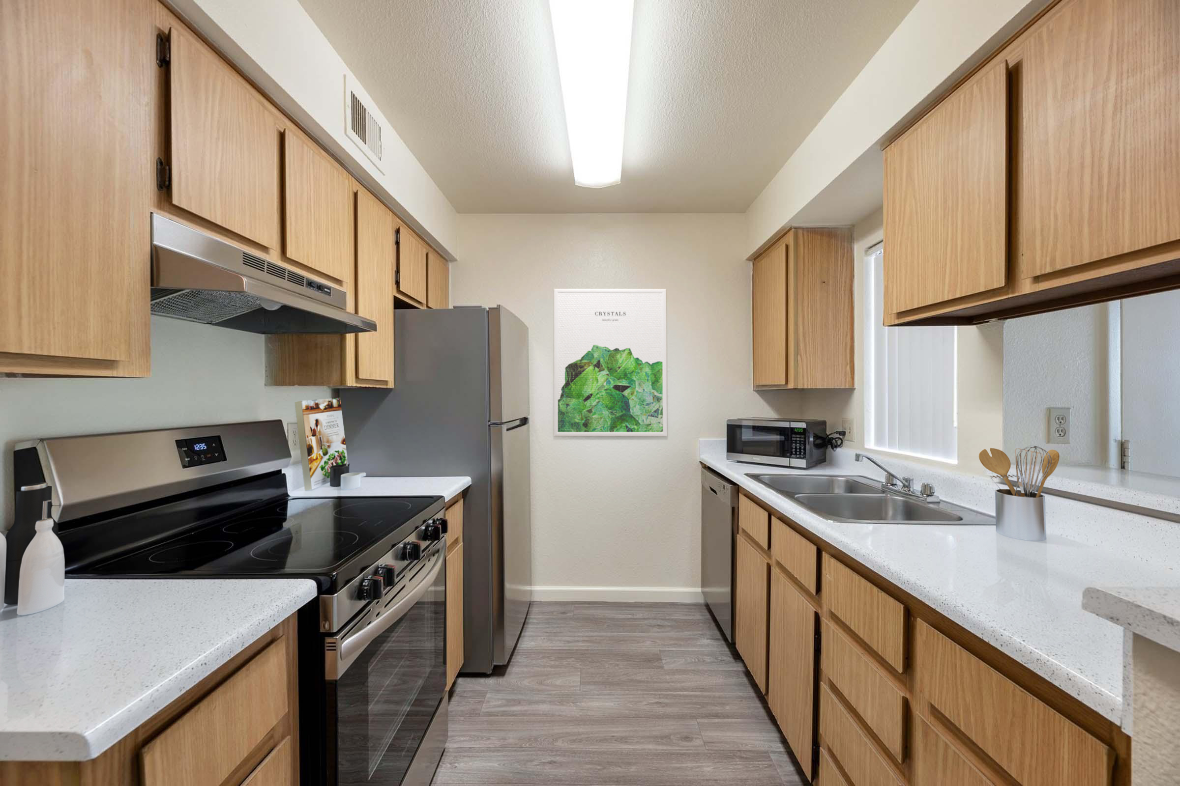 A modern kitchen featuring wooden cabinets, stainless steel appliances, and a white countertop. The space includes a stove, refrigerator, microwave, and a double sink. A framed artwork of green leaves is displayed on the wall, adding a decorative touch to the bright, well-lit area.