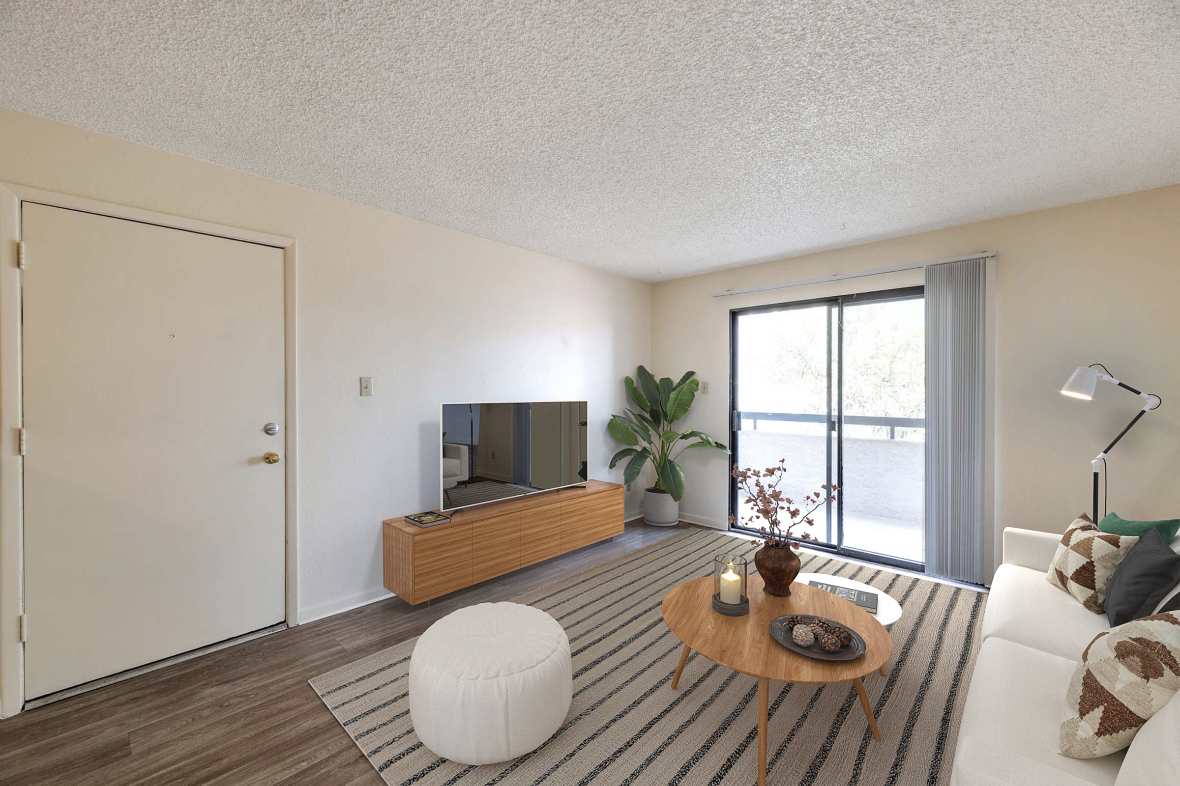 A modern living room featuring a white sofa with decorative cushions, a round coffee table, and a large flat-screen TV mounted on a wooden stand. There’s a green potted plant and a soft pouf on a striped rug. A sliding glass door leads to a balcony, letting in natural light.