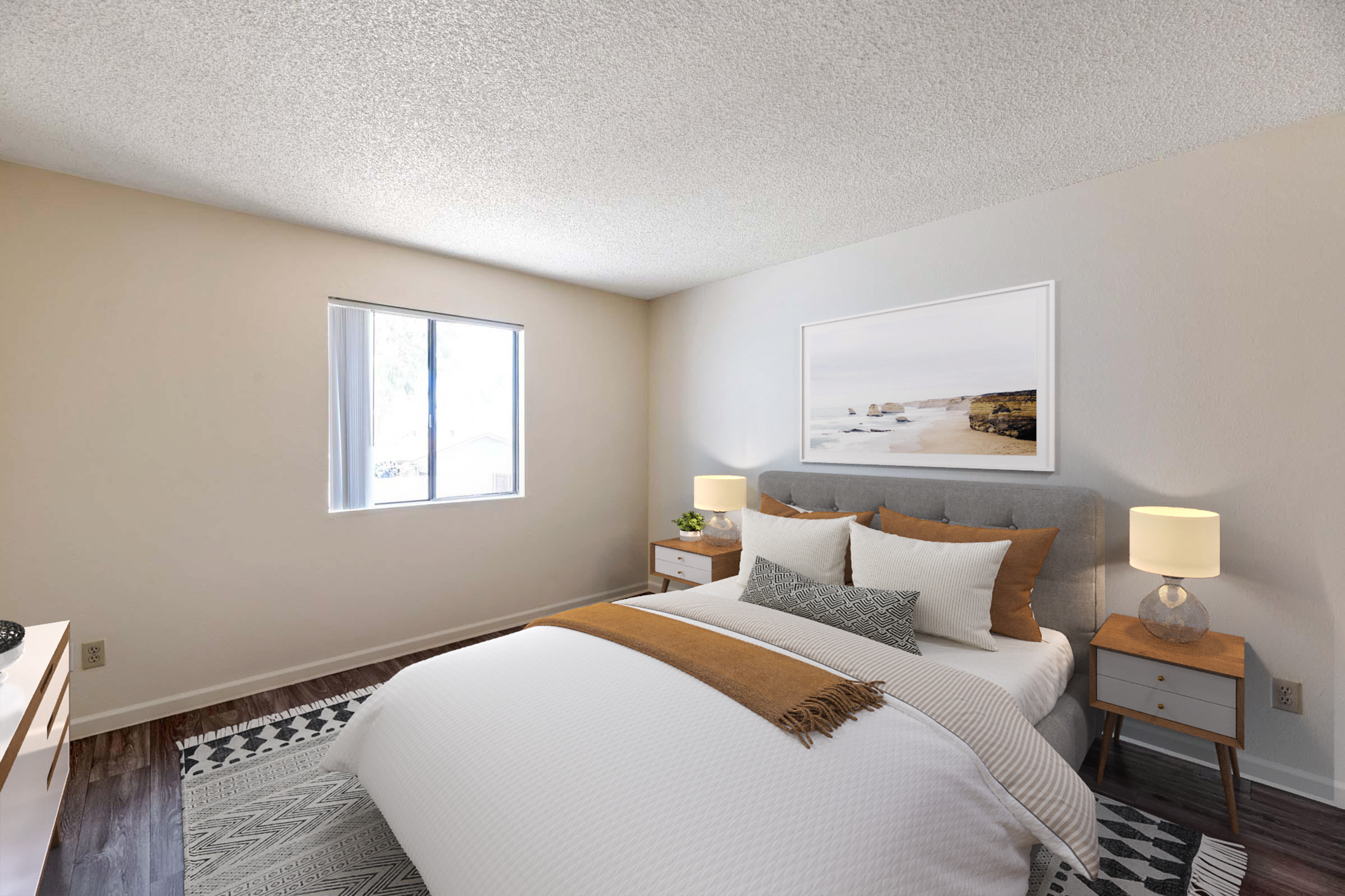 A cozy bedroom featuring a grey upholstered bed with white and beige bedding. There are two nightstands with lamps on either side, and a minimalist wall art piece above the bed. A window lets in natural light, and the floor is covered with a patterned rug, creating a warm and inviting atmosphere.