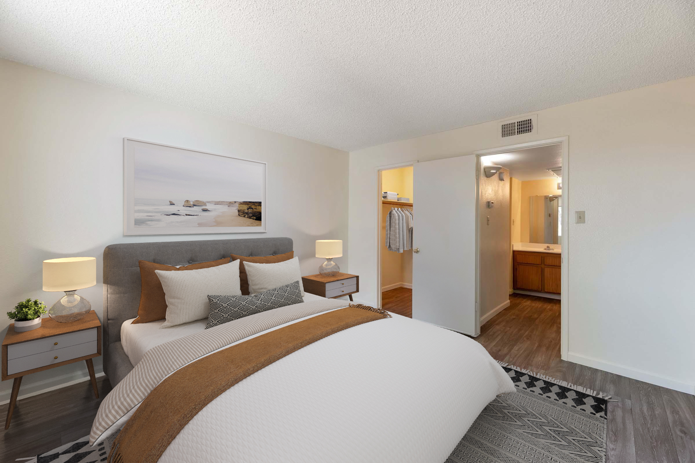 A cozy bedroom featuring a gray upholstered bed with white and brown bedding. There are two bedside tables with lamps and a plant. A large framed picture of a coastal scene is on the wall. An open doorway leads to a bathroom, visible in the background, and a closet is also accessible. The room has neutral walls and hardwood flooring.