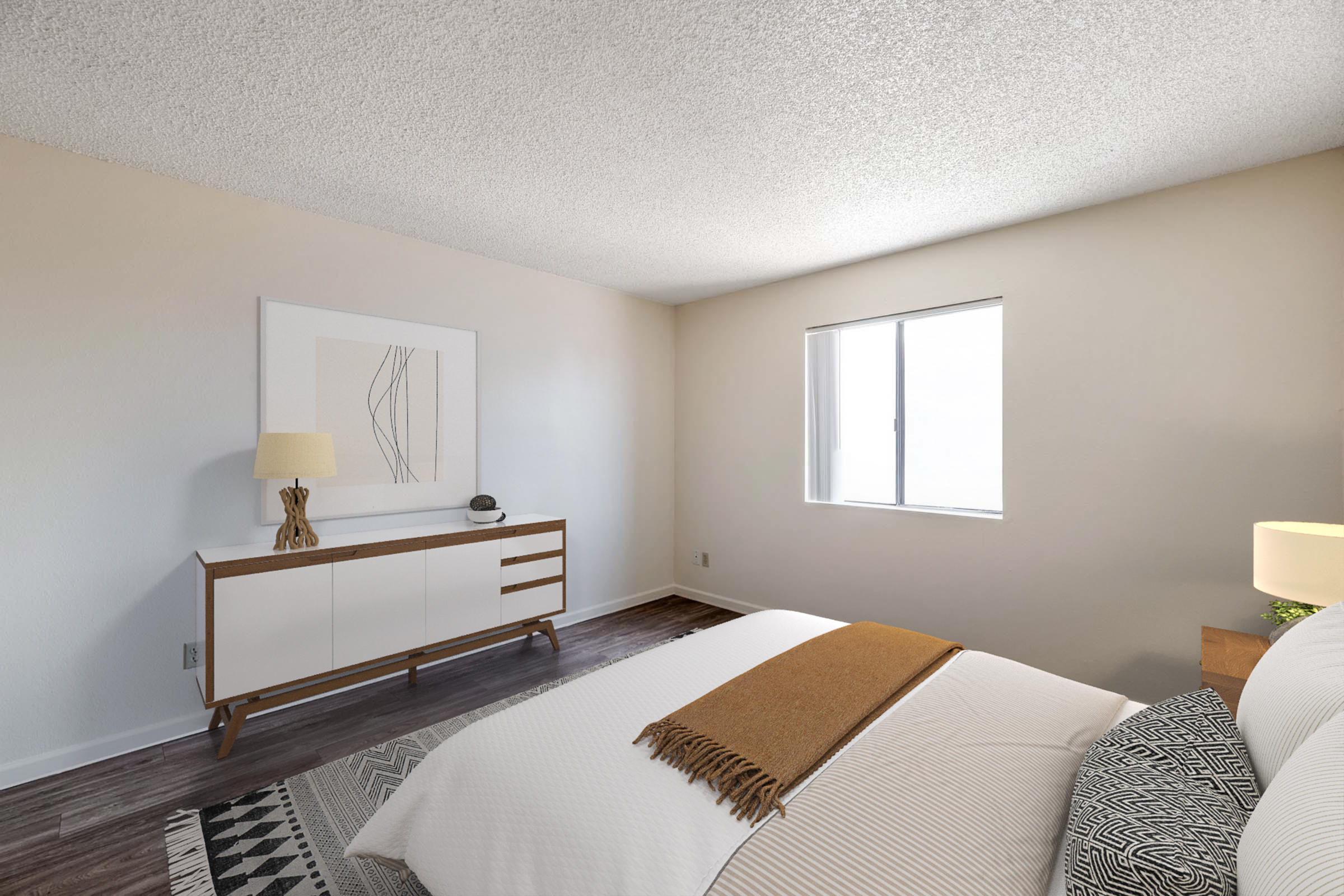 A well-lit bedroom featuring a cozy bed with white linens and a brown throw blanket. There's a stylish dresser with a lamp and decorative items, along with a window allowing natural light. The walls are a soft color, and the floor has a textured rug, creating a warm and inviting atmosphere.