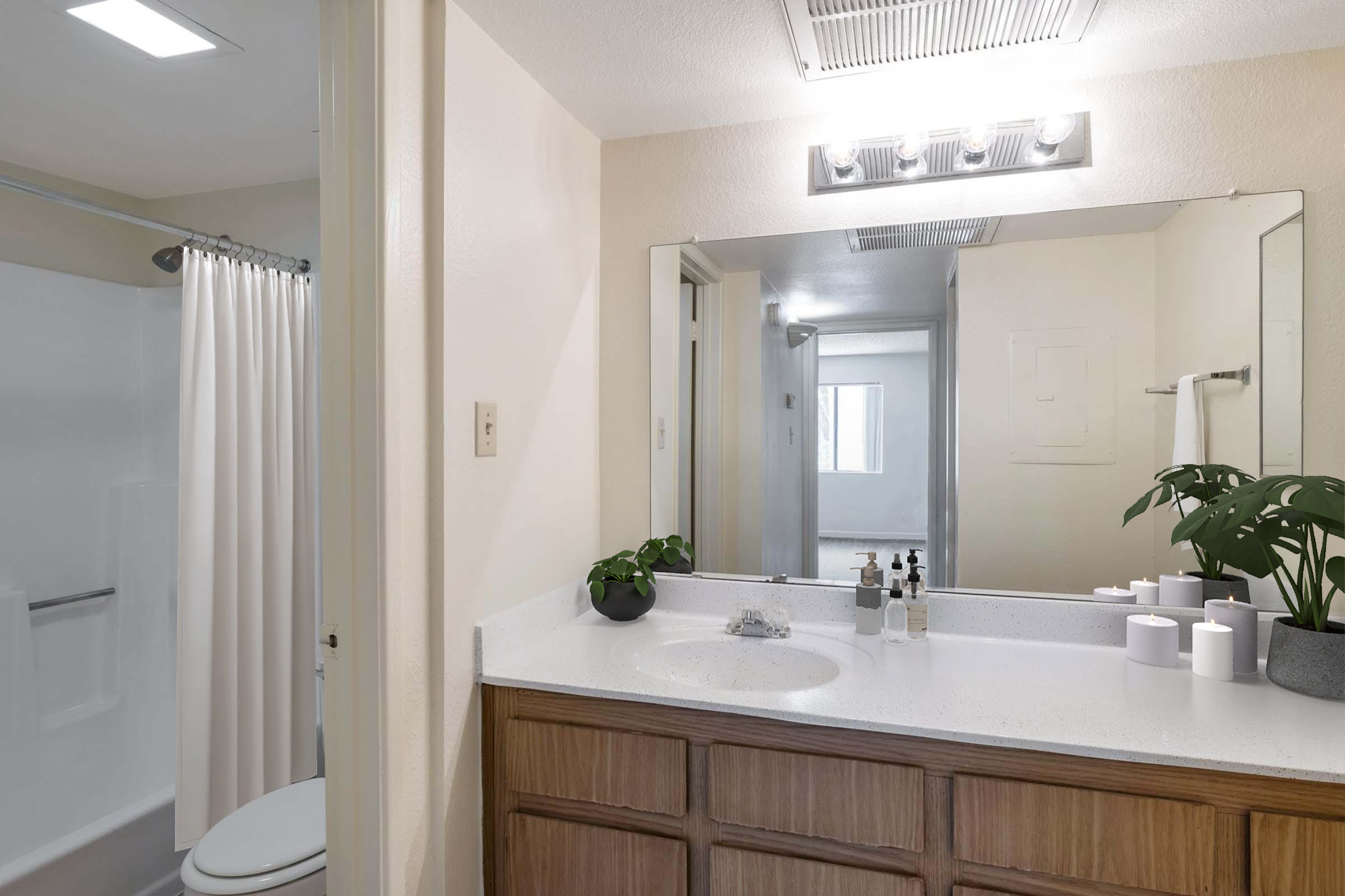 A modern bathroom featuring a white countertop with toiletries, a large mirror, and a light fixture above. To the left, there's a shower with a curtain, and a potted plant adds a touch of greenery. The space is well-lit, creating a clean and inviting atmosphere.