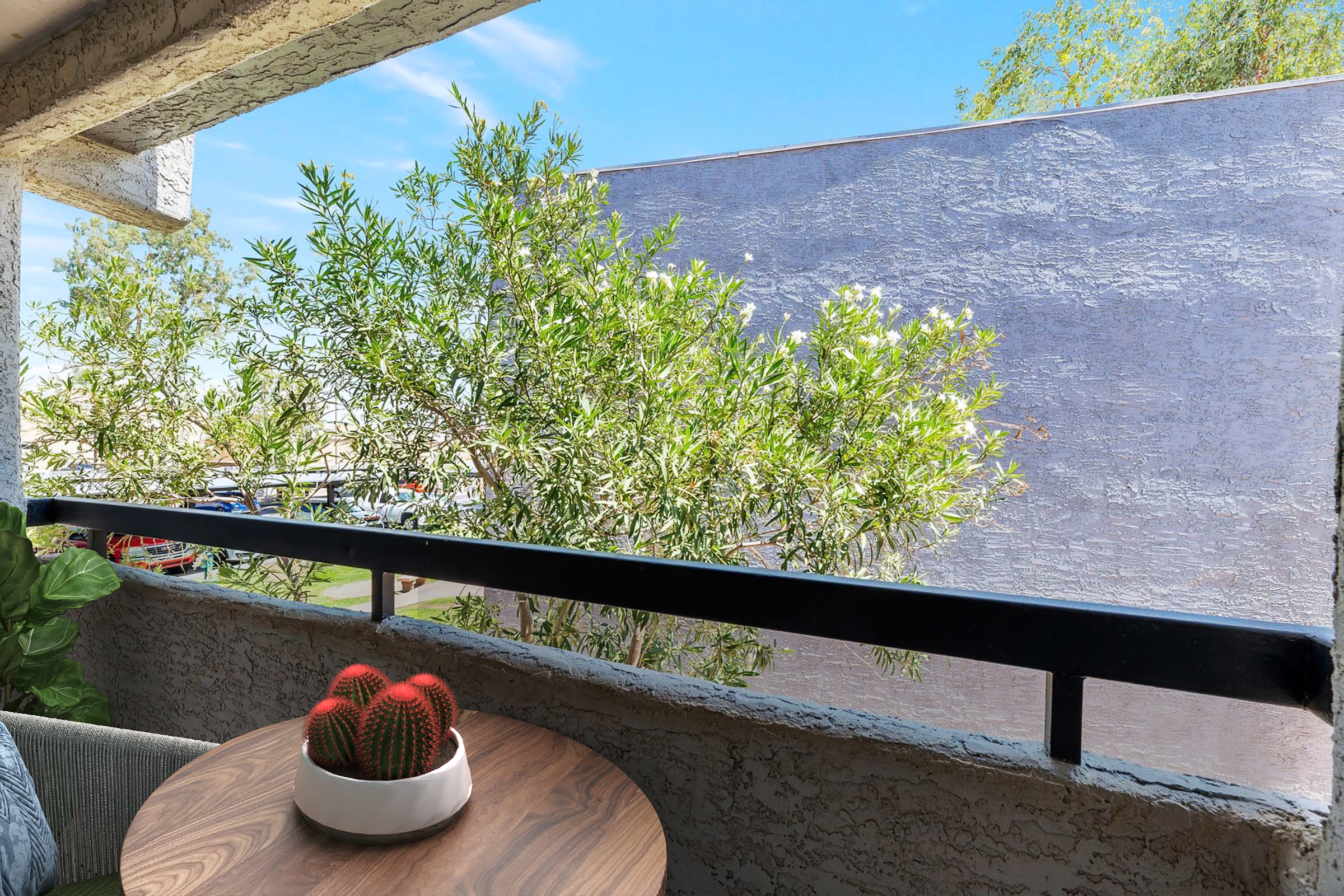 A cozy balcony scene featuring a small wooden table with a potted cactus. In the background, green shrubs and a textured wall are visible under a clear blue sky, creating a relaxed outdoor atmosphere.