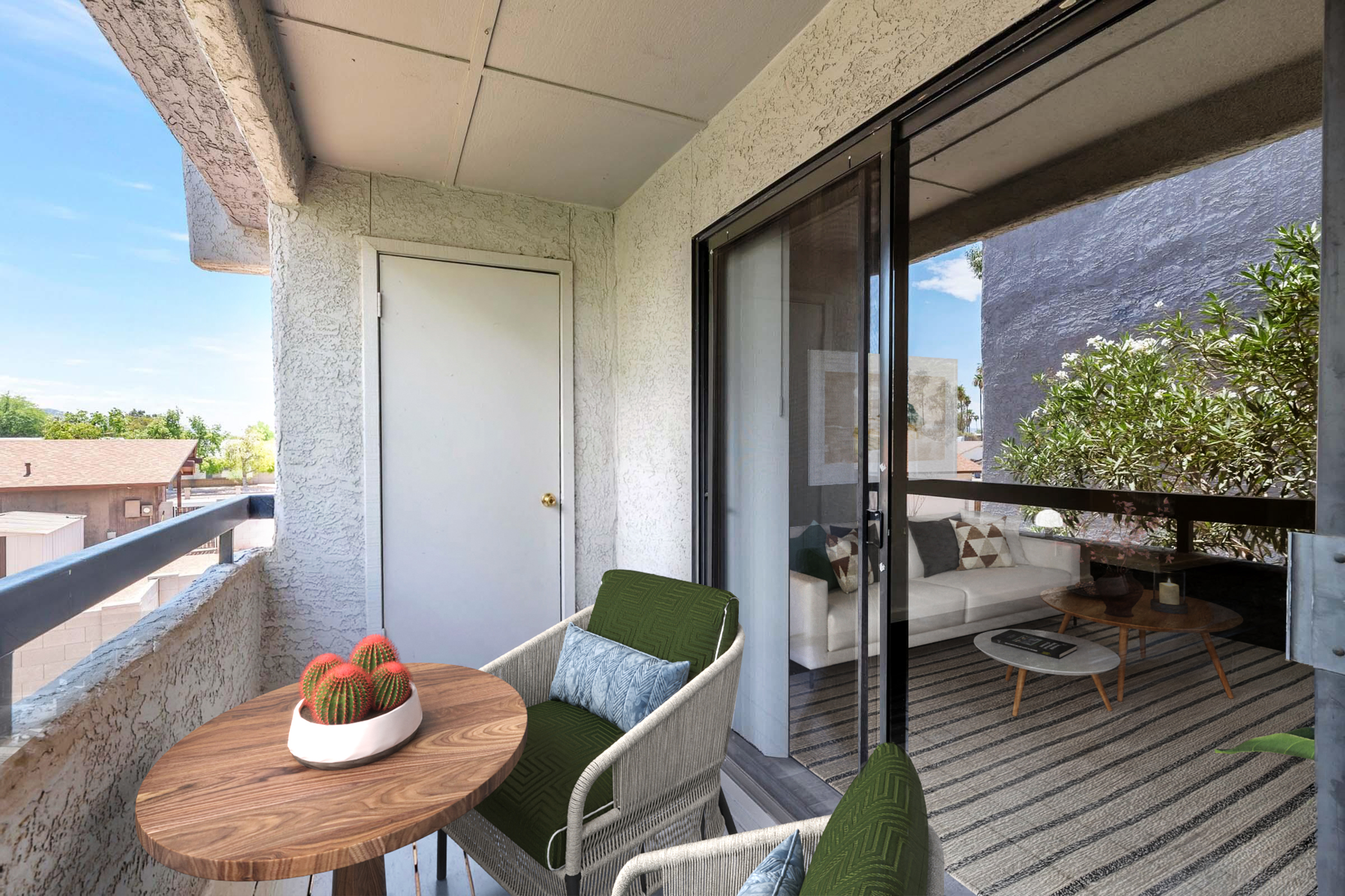 A cozy balcony featuring a small wooden table with a potted cactus arrangement, two comfortable chairs with green cushions, and a partially open sliding glass door leading to a living area. The balcony has light-colored walls and a view of surrounding buildings and greenery under a clear blue sky.