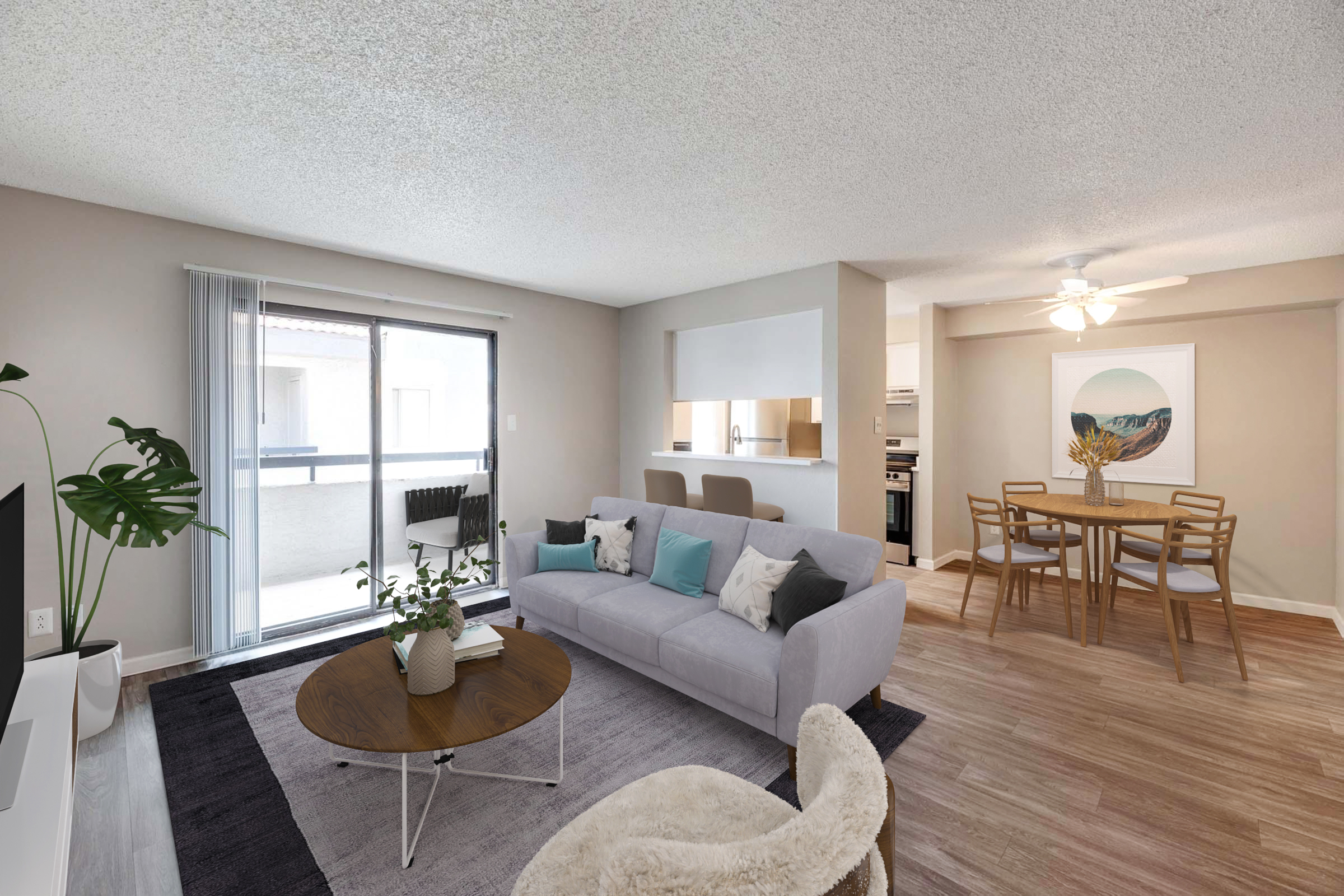 A bright, modern living room featuring a light gray sofa with decorative pillows, a round coffee table, and a large indoor plant. The space opens to a balcony through sliding glass doors. In the background, a dining area with a wooden table and chairs, adjacent to a well-lit kitchen.
