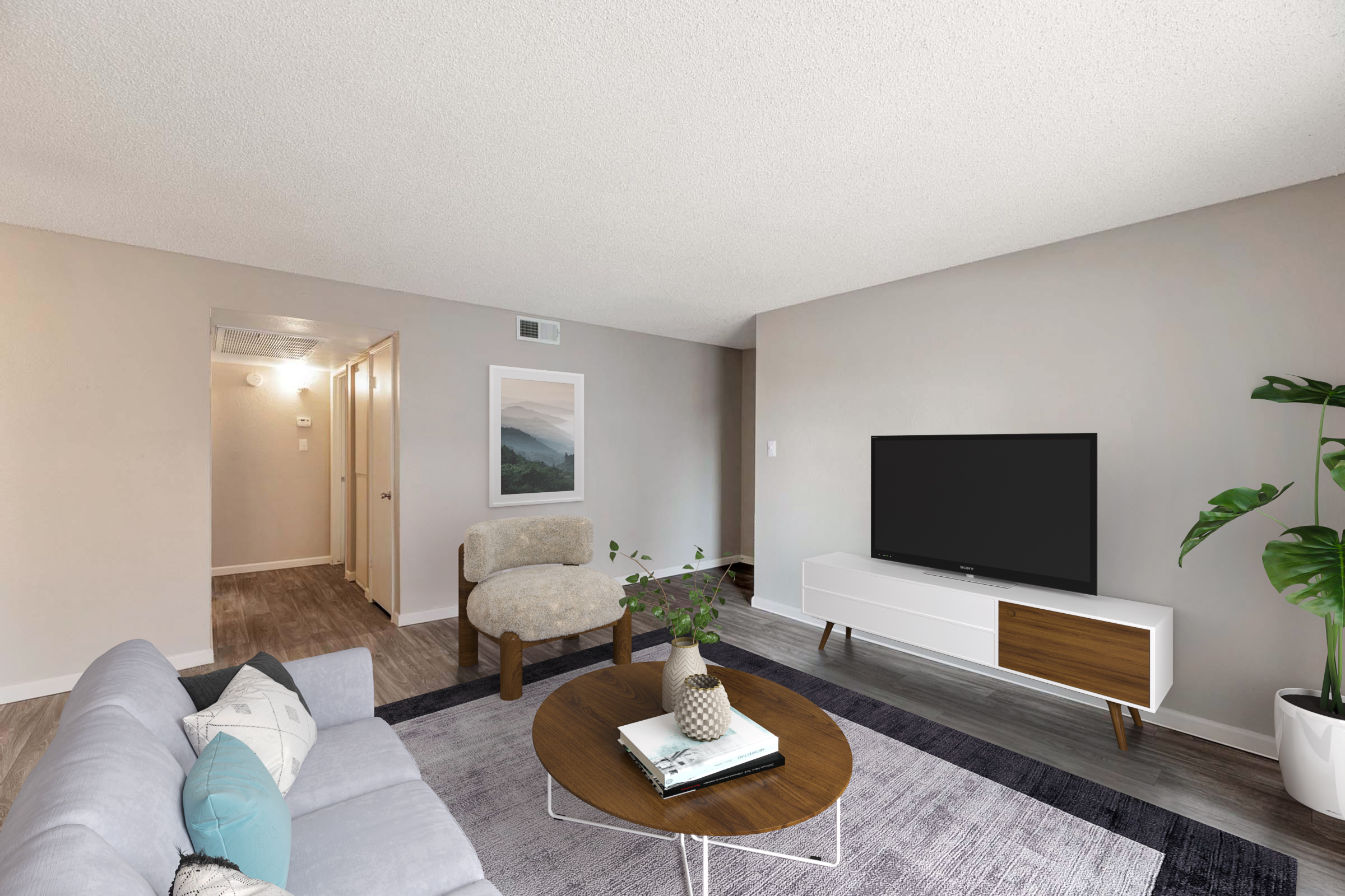 A modern living room featuring a light gray sofa with decorative pillows, a round wooden coffee table, and a flat-screen TV on a white media console. A potted plant adds a touch of greenery, and there are light-colored walls and flooring. A hallway can be seen in the background.