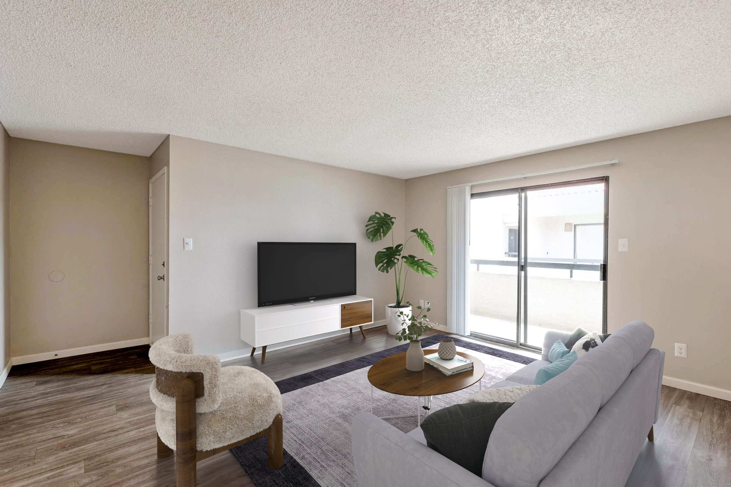 A modern living room featuring a light gray sofa, a round wooden coffee table with a plant, and a cozy chair. A flat-screen TV is mounted on the wall above a white TV stand. The room has neutral-colored walls and a sliding glass door that opens to a balcony, allowing natural light to fill the space.