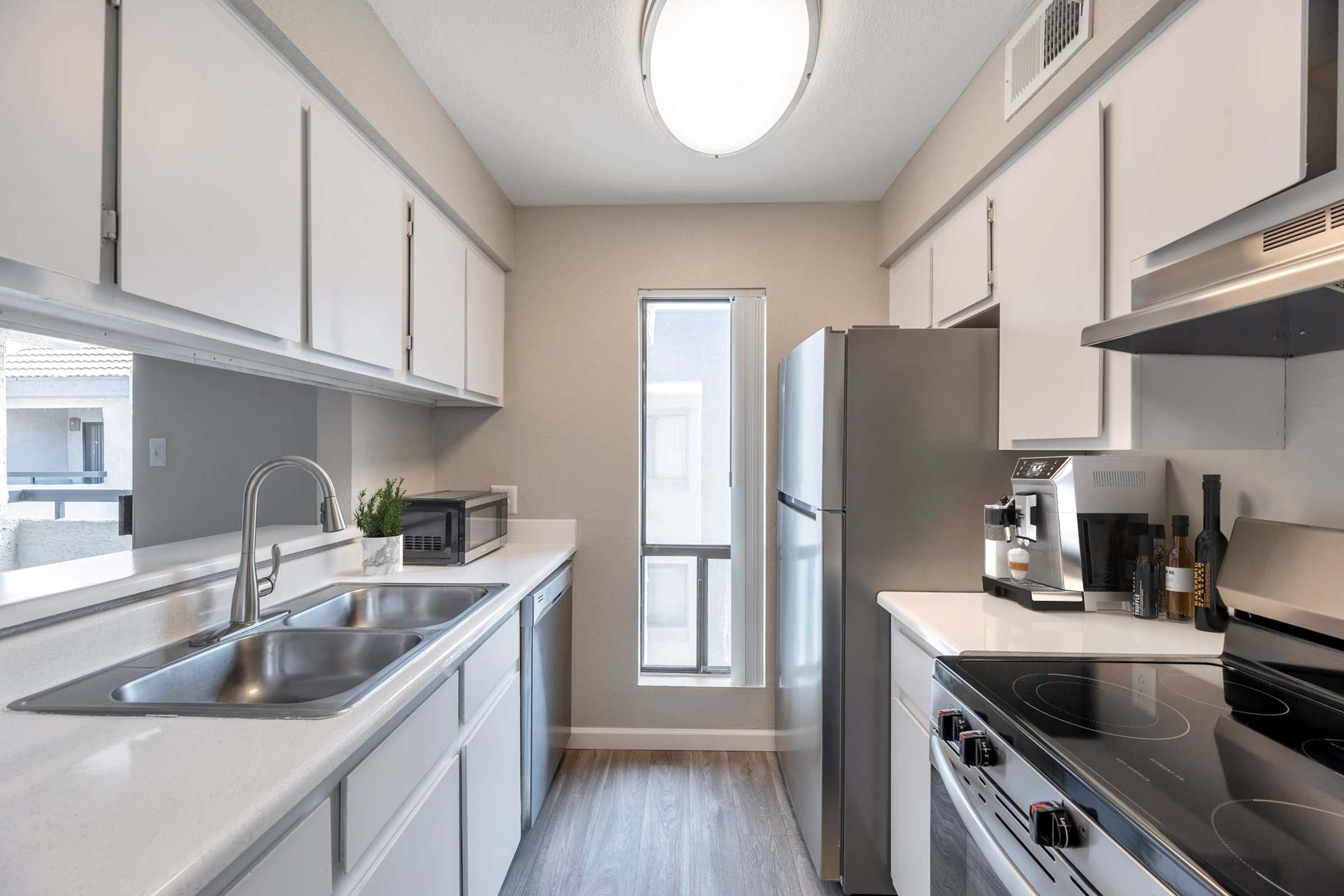 A modern kitchen featuring white cabinetry, a double sink, and stainless steel appliances including a refrigerator and stove. Natural light enters through a window, illuminating the countertop. A coffee maker and wine bottles are visible, adding to the kitchen's functionality and style.