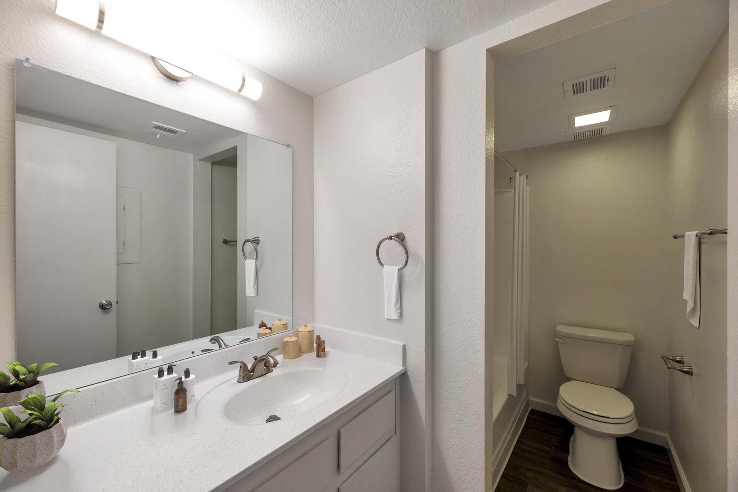 A modern bathroom featuring a double sink vanity with a large mirror, accompanied by a plant decoration. To the right, a small enclosed shower area is visible, alongside a toilet. The walls are painted in neutral tones, creating a bright and clean atmosphere, illuminated by overhead lighting.