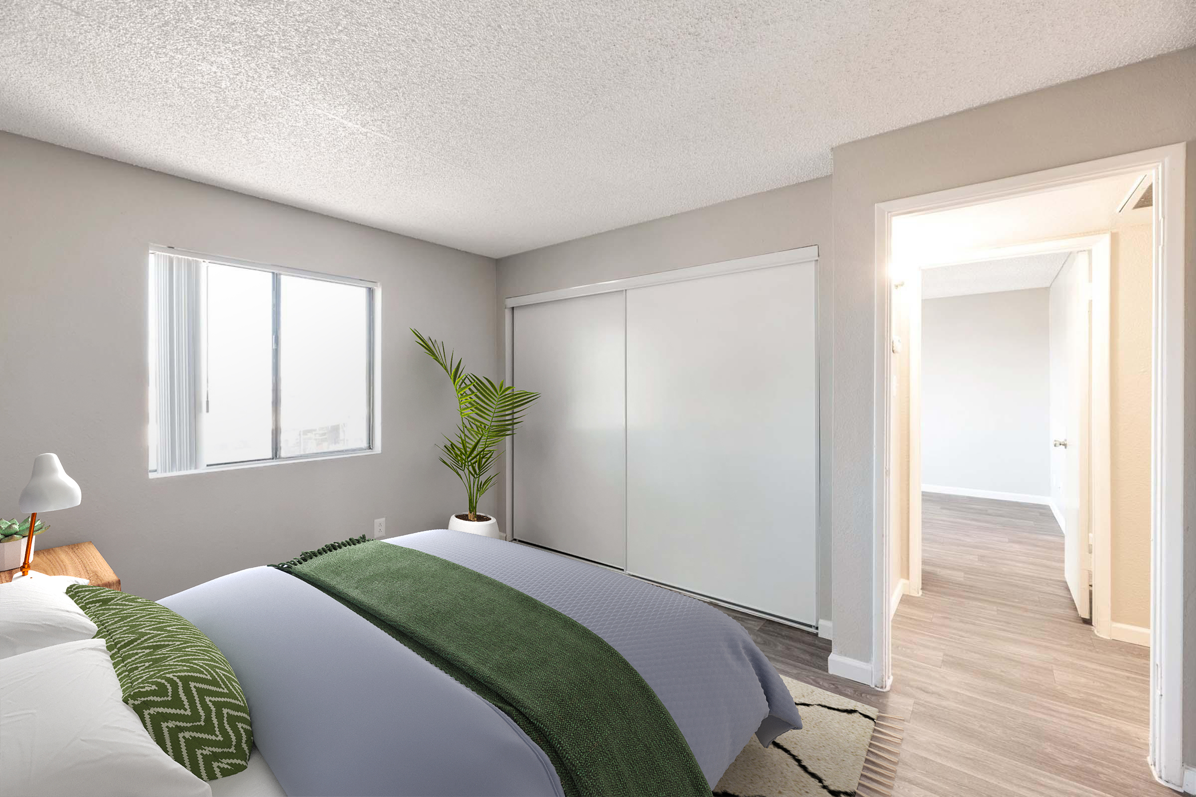 A well-lit bedroom featuring a bed with a gray and green duvet, a decorative plant beside a bedside table, and a sliding door wardrobe. The room has light gray walls and a doorway leading to another space, highlighting a cozy and inviting atmosphere.