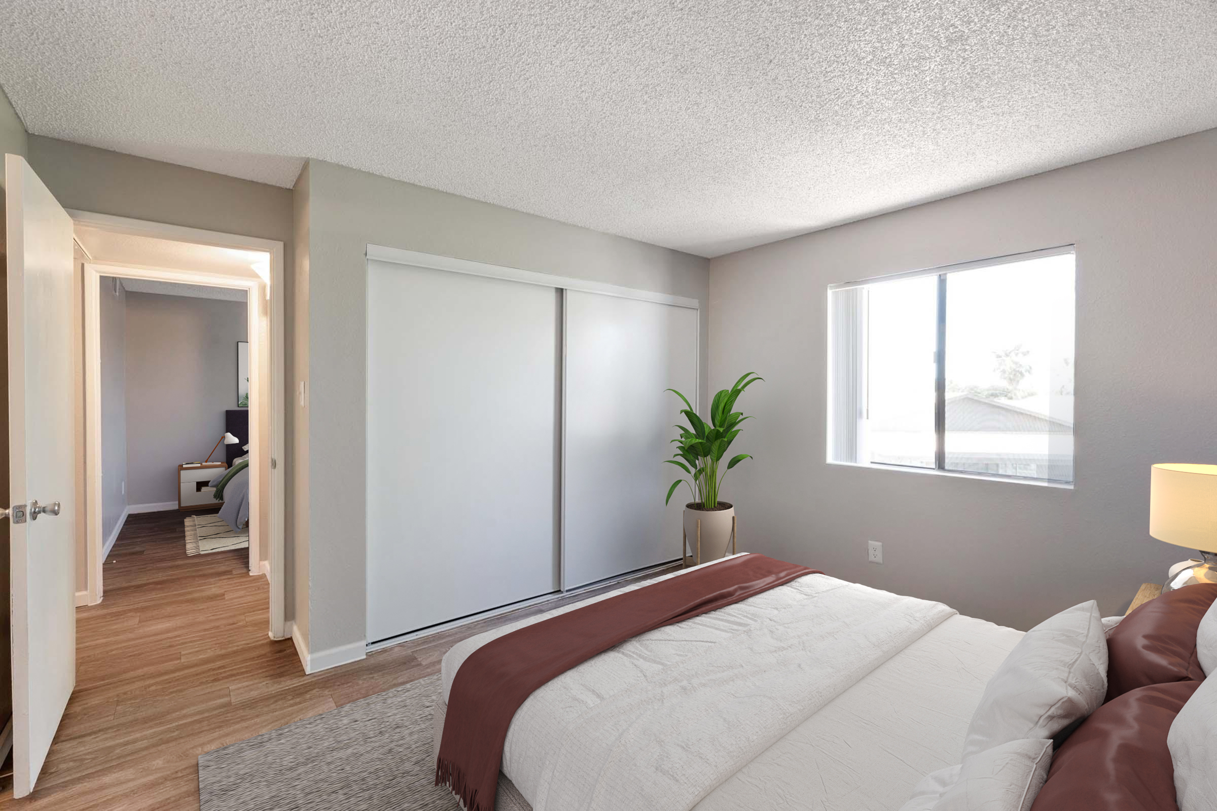 A cozy bedroom featuring a bed with white linens and a brown throw blanket. There is a potted plant near the window, providing natural light. A closet with sliding doors is visible, along with an open doorway leading to another room, creating a welcoming and bright atmosphere.