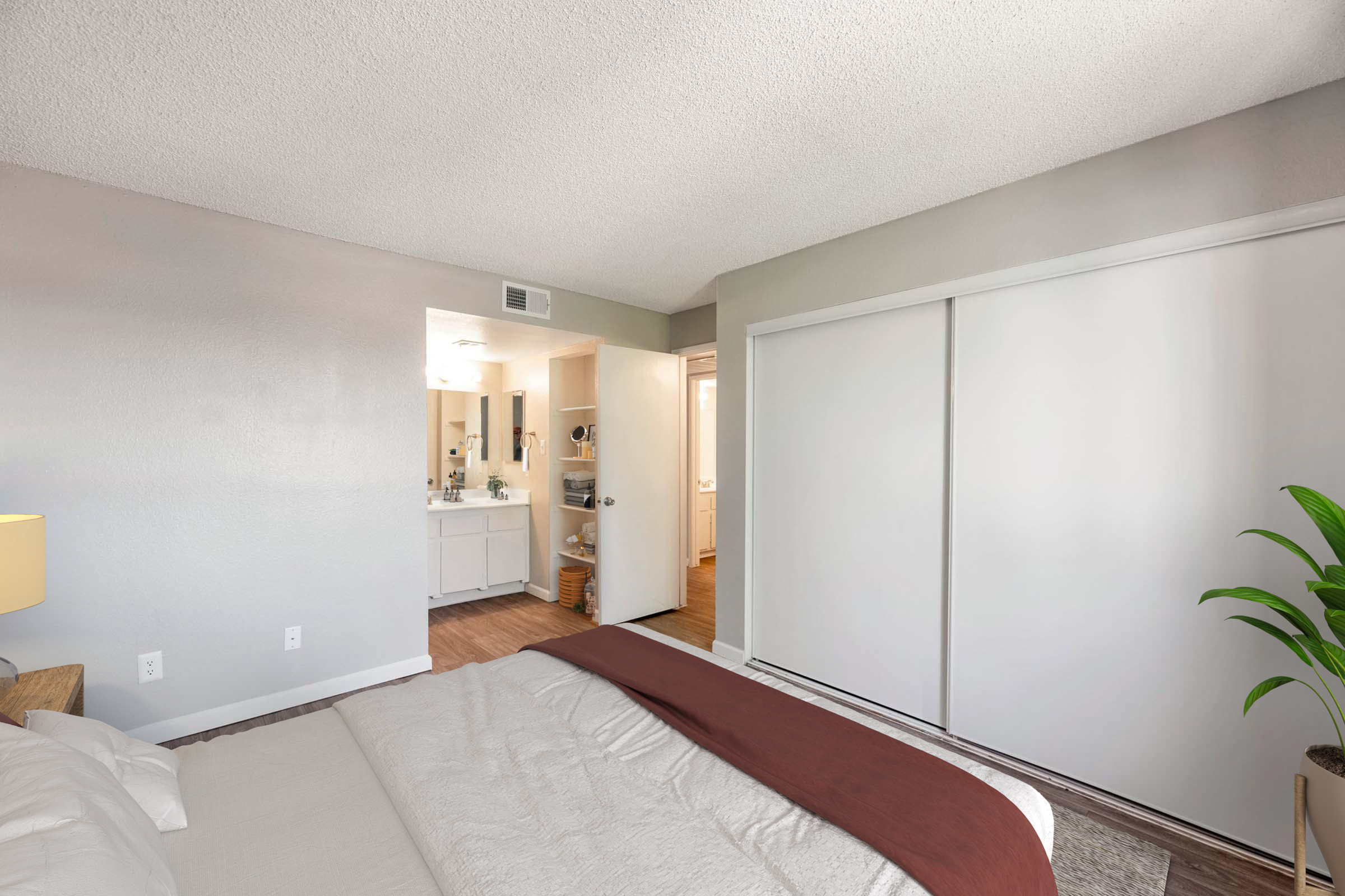 A well-lit bedroom featuring a bed with a white and maroon comforter. There is a bedside lamp on a wooden nightstand. In the background, an open door leads to a bathroom area, showcasing a sink and white cabinetry. A tall green plant adds a touch of greenery to the room. The walls are painted a light gray.