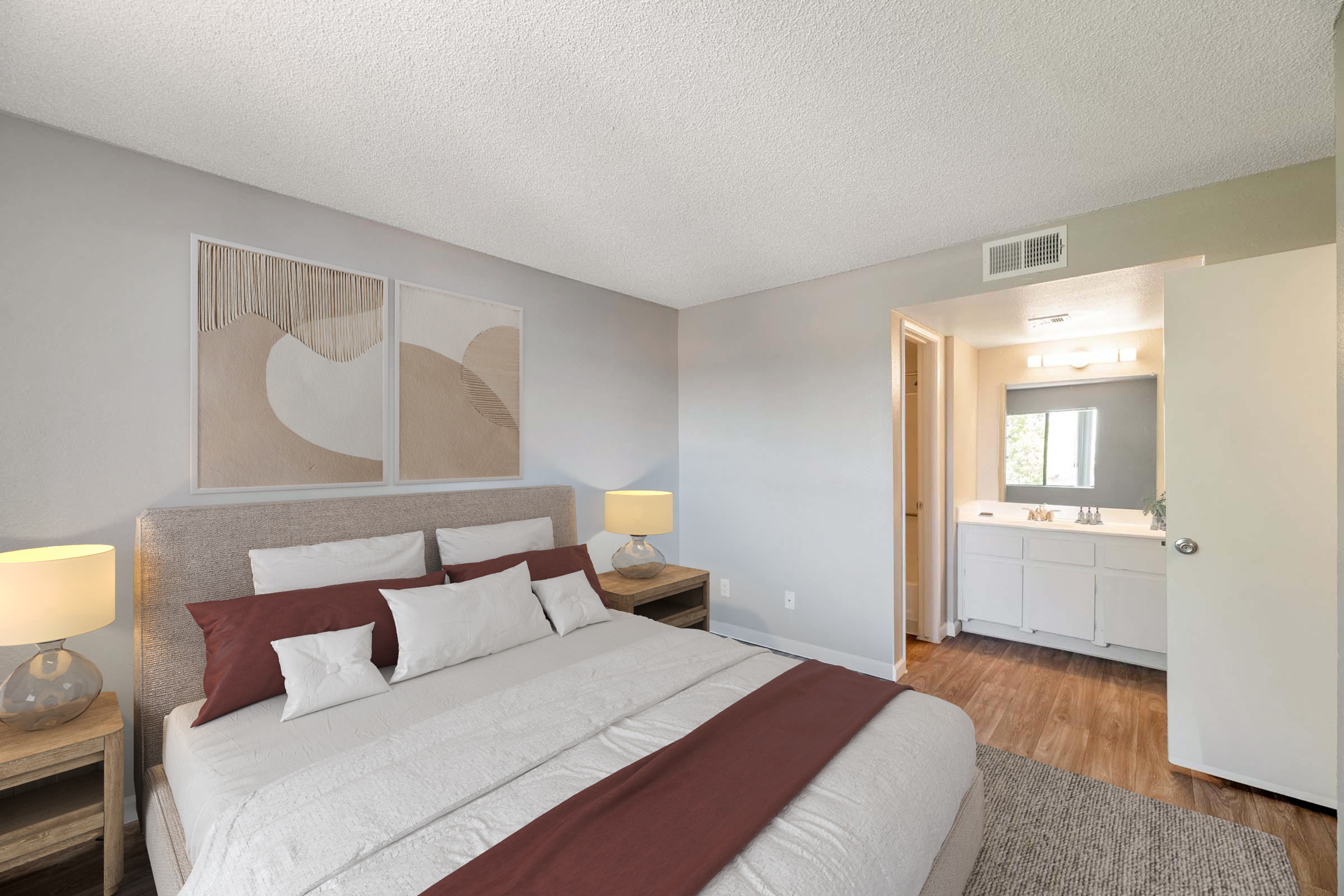 A cozy bedroom featuring a large bed with white and burgundy bedding, bedside tables with lamps, and abstract artwork on the wall. An open door leads to a bathroom area with a large mirror and vanity. The room has a neutral color scheme and wooden flooring, creating a warm and inviting atmosphere.