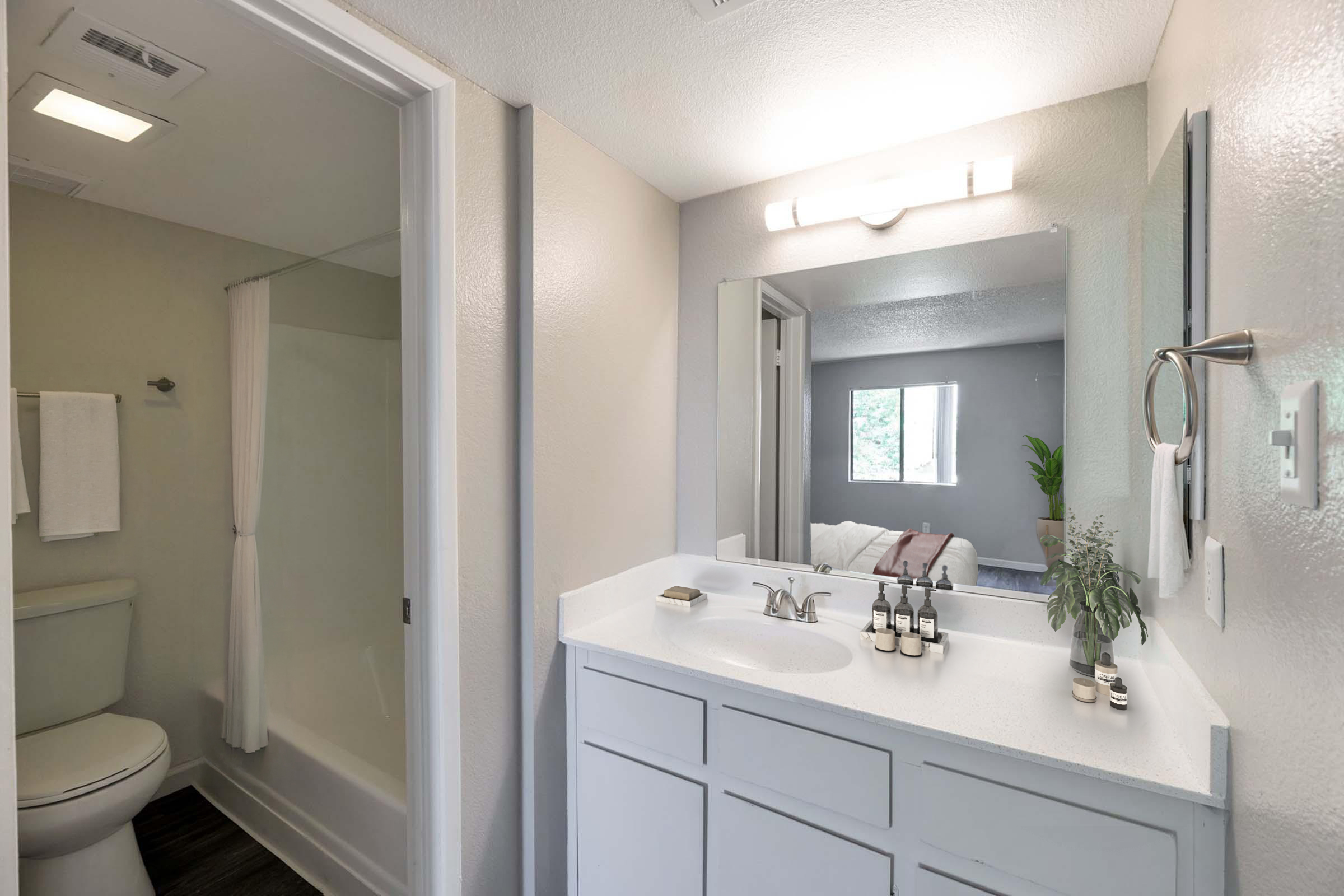 A modern bathroom featuring a white vanity with a mirror, a sink with soap dispensers, and neatly arranged toiletries. A shower area is visible with a frosted glass door. The walls are painted light gray, and a window in the background offers a view of greenery. Towels hang neatly on the side.