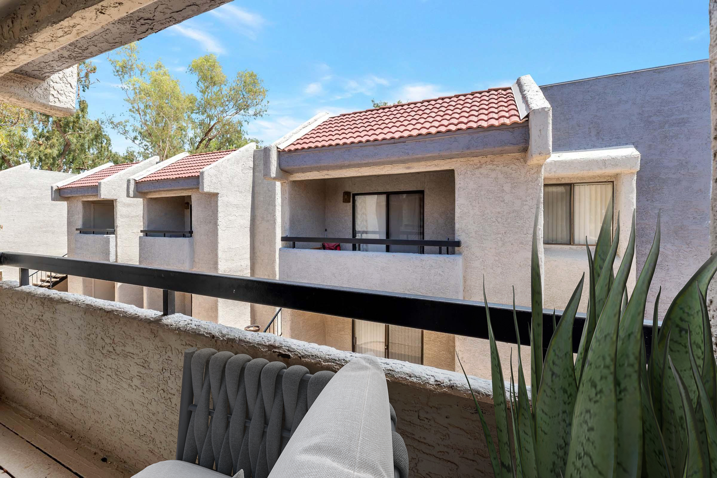 A view from a balcony showing two buildings with tiled roofs. The buildings have multiple windows and balconies. In the foreground, there is a gray chair and green plants. The sky is blue with a few clouds, and the trees in the background add greenery to the scene.