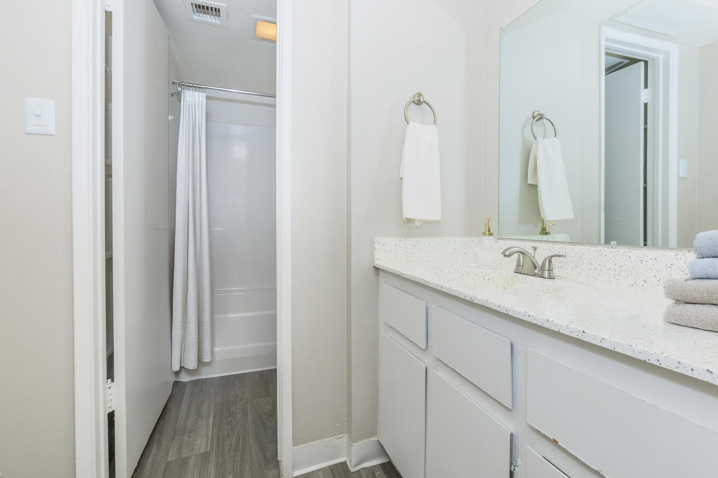 A clean, modern bathroom featuring a white countertop with neatly stacked towels, a shower area with a white curtain, and a large mirror. The walls are painted in a neutral color, and the floor has a wood-like finish.