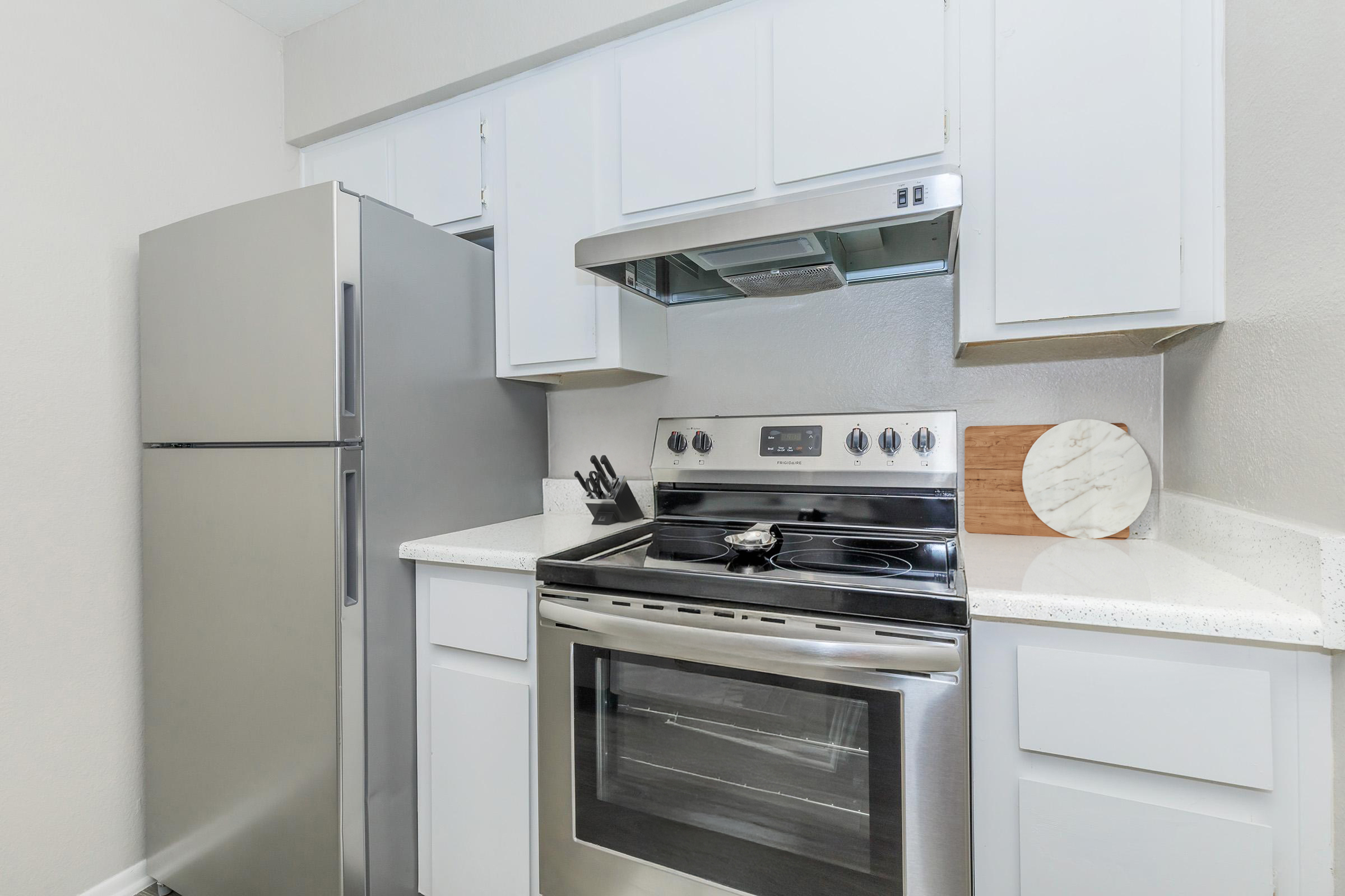 A modern kitchen featuring stainless steel appliances, including a refrigerator and an oven with a cooktop. The cabinets are white, and the countertops are a light, stone-like material. A wooden cutting board and utensils are neatly arranged on the countertop, contributing to the clean and organized appearance.