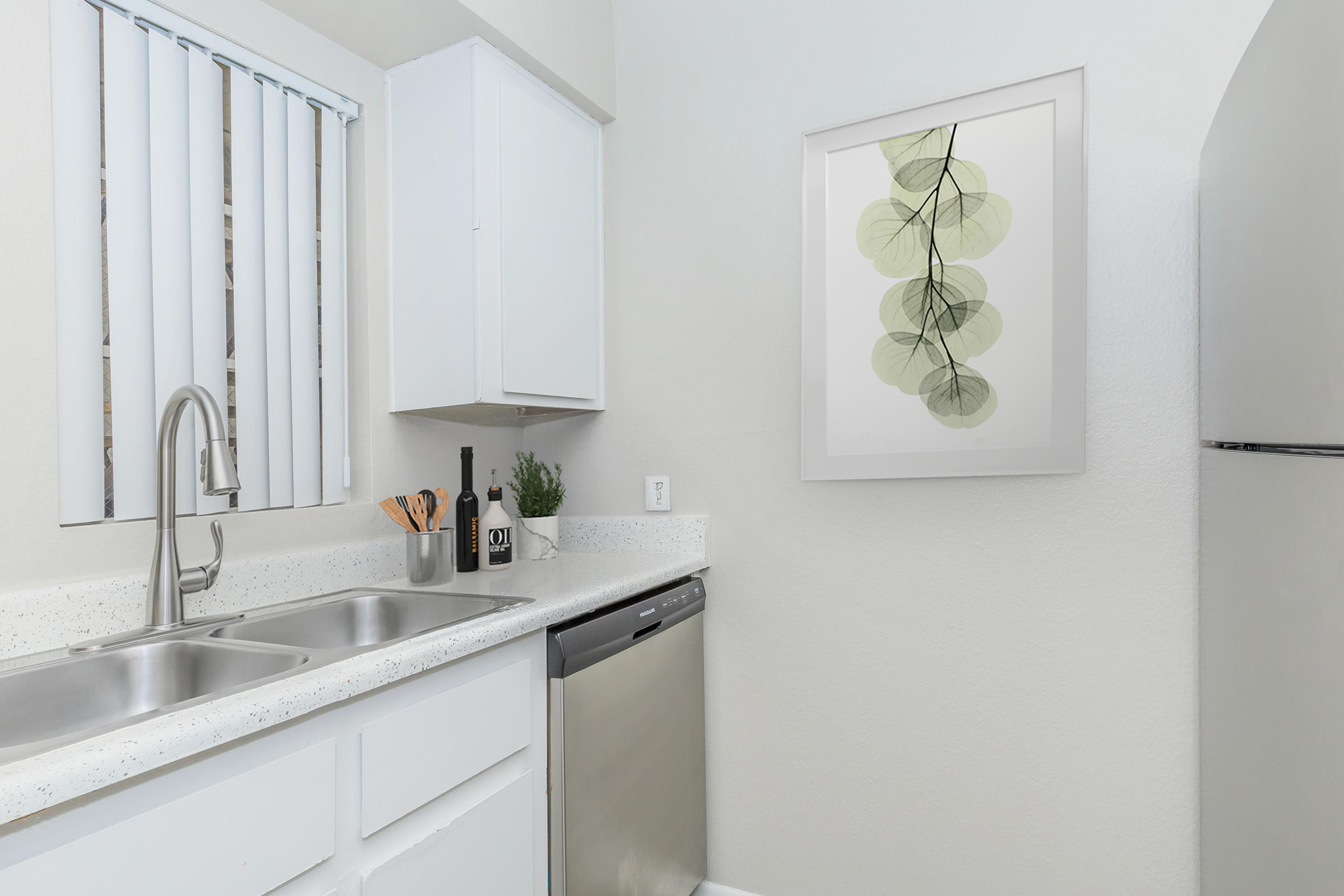 A modern kitchen featuring white cabinetry, a silver dishwasher, a double sink, and a stainless steel refrigerator. The countertop is adorned with kitchen utensils and a decorative plant. A framed piece of art with green leaves is displayed on the wall above the sink. Natural light streams in through vertical blinds.