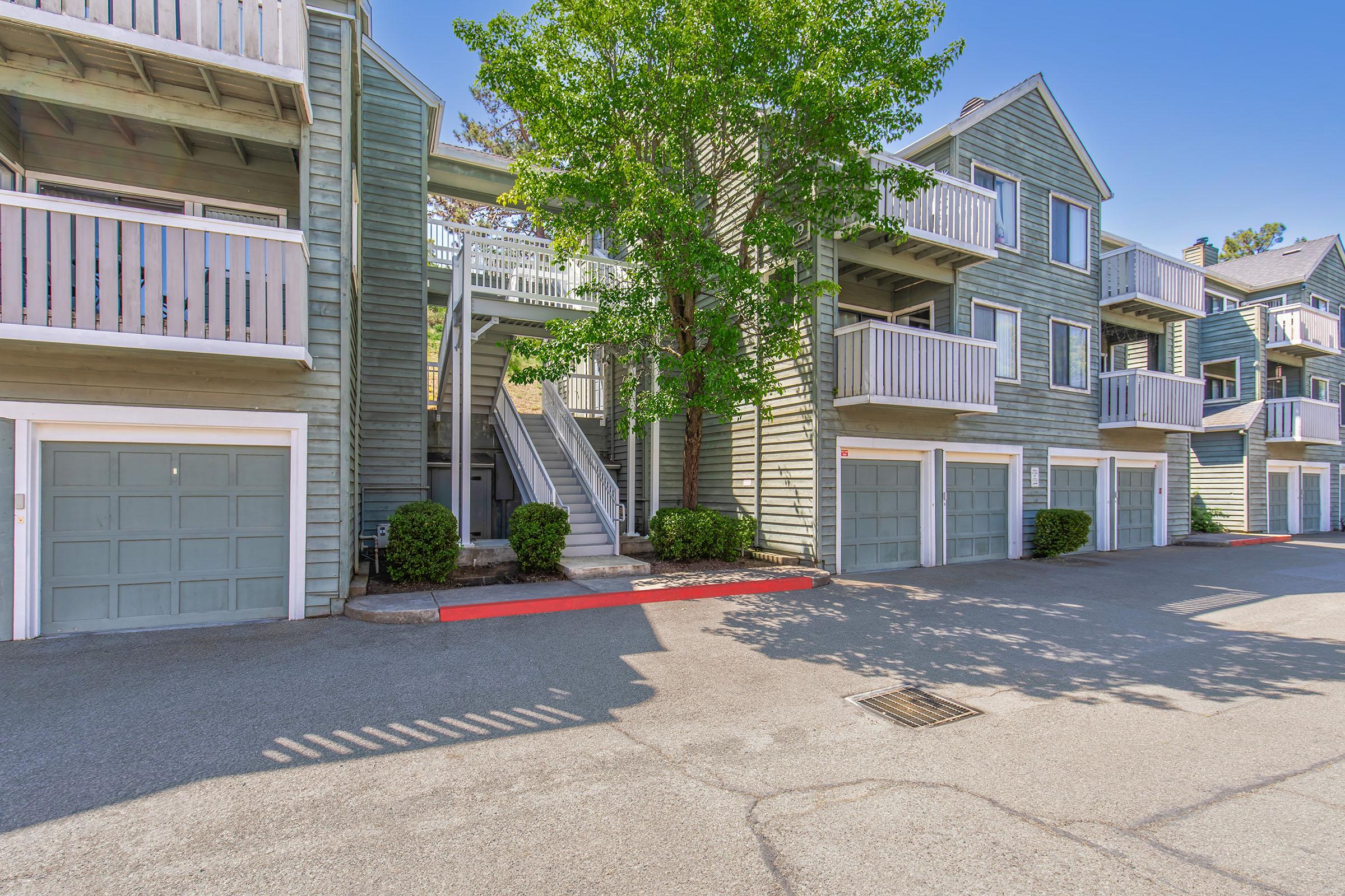 A multi-story residential building with green wooden siding, featuring balconies on the second floor. There are garages on the ground level, and neatly trimmed bushes line the pathway. A large tree provides shade in front of the building, and the parking area is paved with visible shadows from the structure.