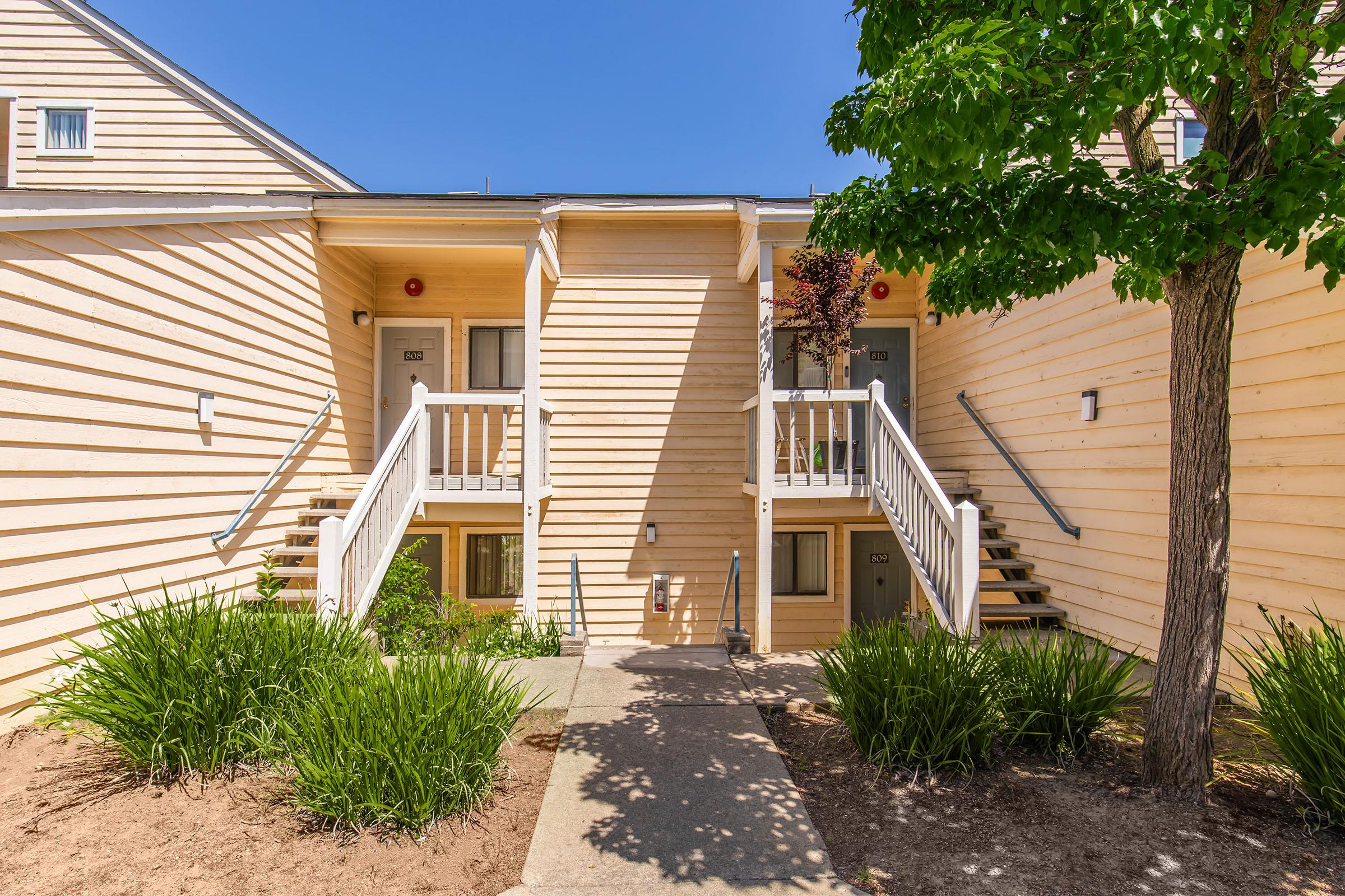 A two-story building with yellowish wooden exterior. There are two sets of white stairs leading to separate doors. The walkway is lined with green plants and grass. A tree is visible on the right side, under a clear blue sky.