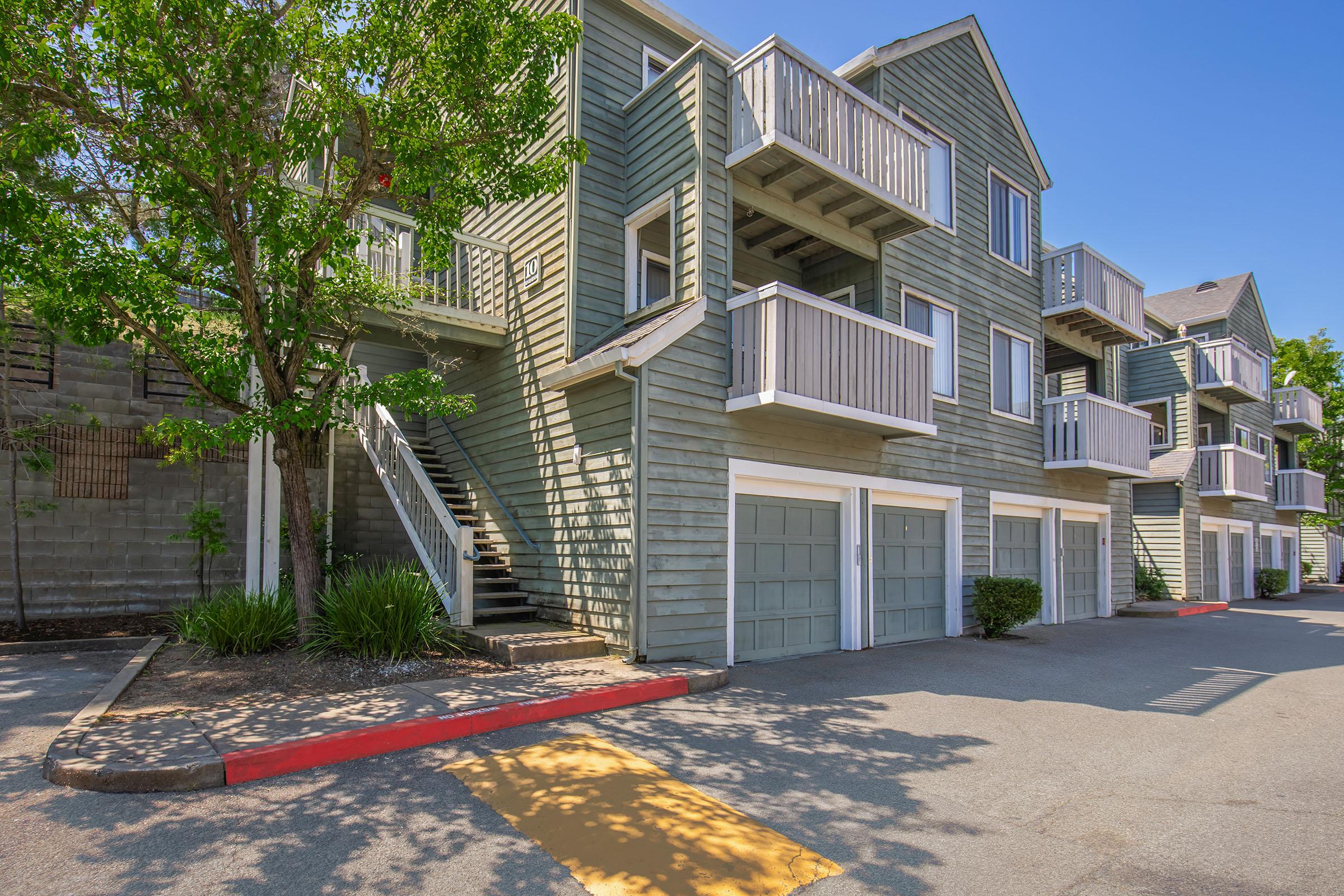A multi-story apartment building with a green wooden exterior, featuring balconies on each unit. The lower level has garages with white doors. A tree and landscaped area are visible in front, along with a paved parking lot and clear blue sky above.