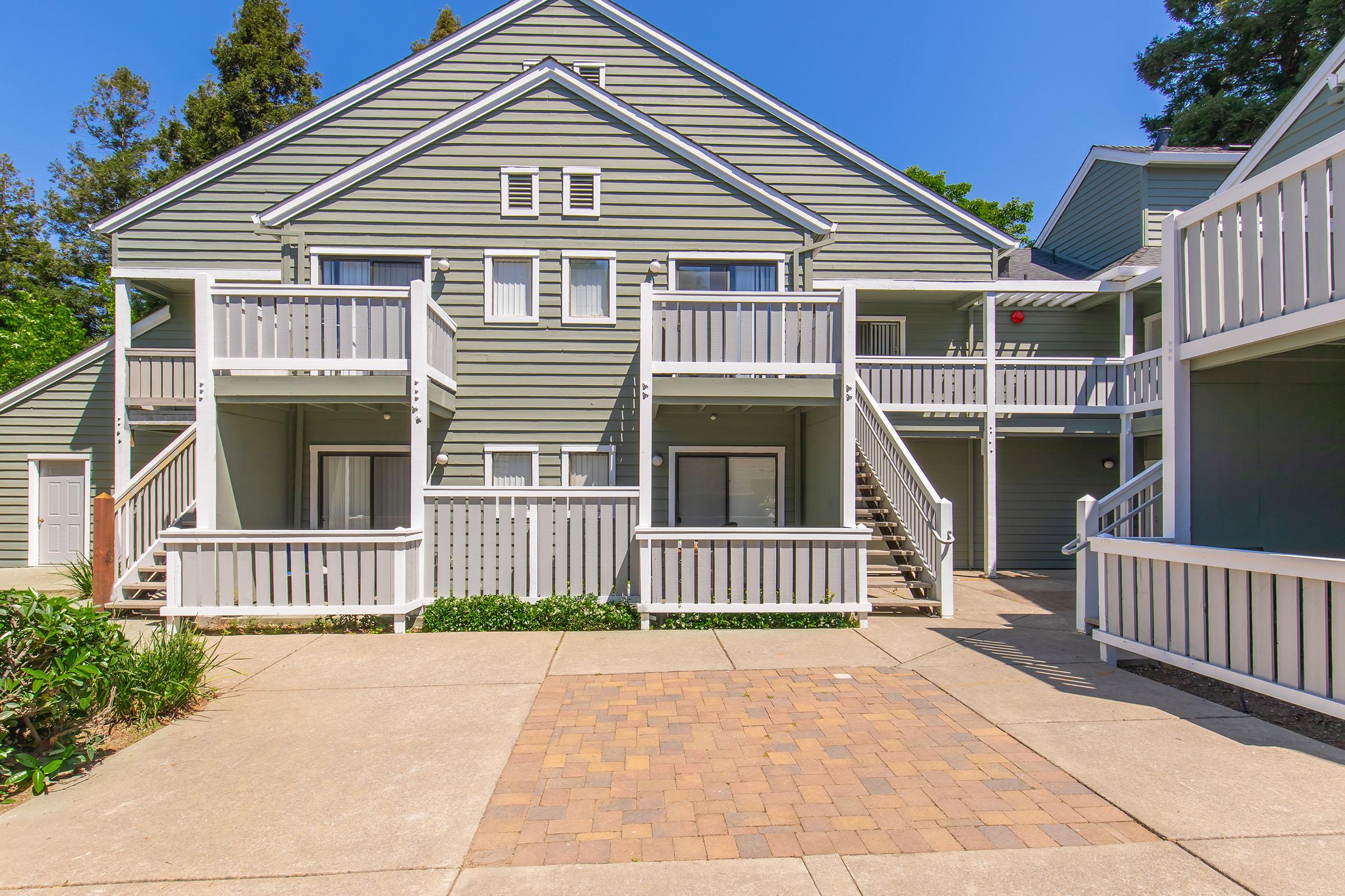 A multi-unit residential building with pale green siding, featuring balconies on the upper levels. The property has a paved walkway in front and is surrounded by shrubs and trees under a clear blue sky. The building consists of two levels with staircases leading to the upper balconies.