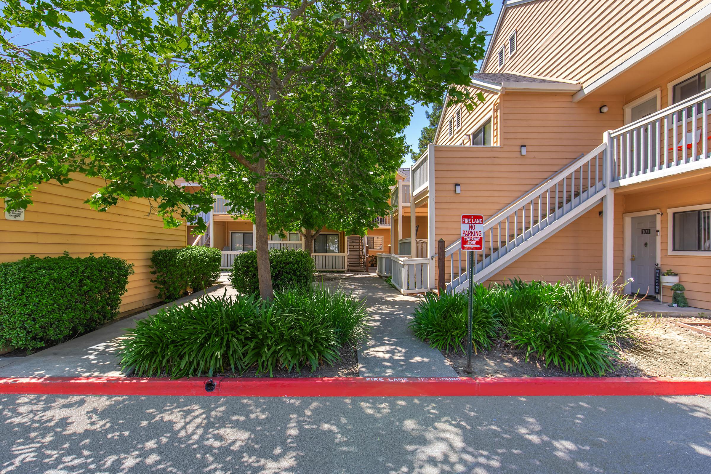 View of a residential complex with two buildings featuring orange siding and white railings. A tree provides shade in the foreground, surrounded by green shrubs. A sign is posted near the pathway, and the ground is paved with a red border. The scene is bright and sunny, indicating a pleasant neighborhood atmosphere.