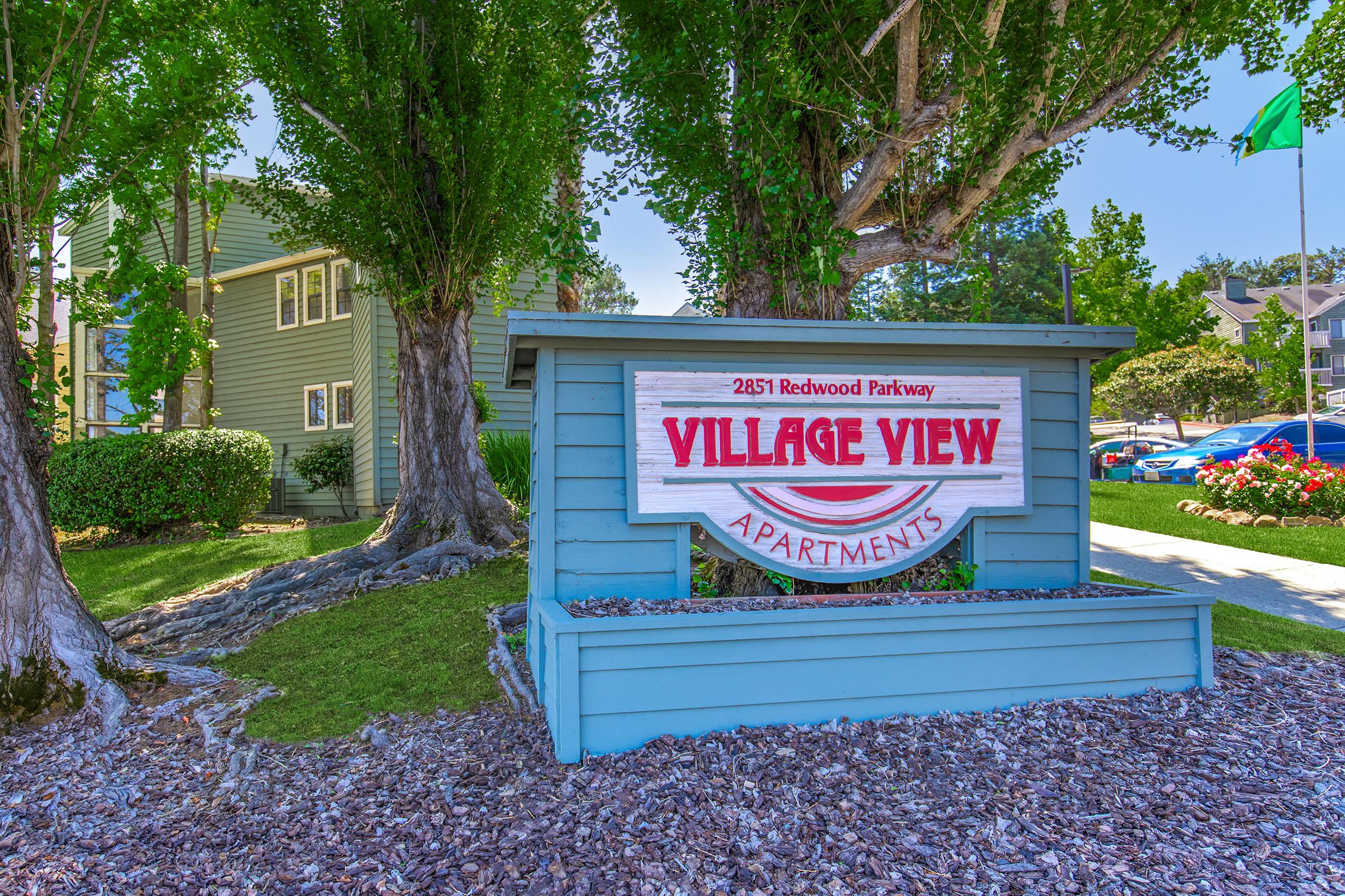 Sign for Village View Apartments located at 2851 Redwood Parkway. The sign features a colorful design, with the name prominently displayed in red letters. Surrounding the sign are green trees and landscaped areas, creating an inviting appearance.
