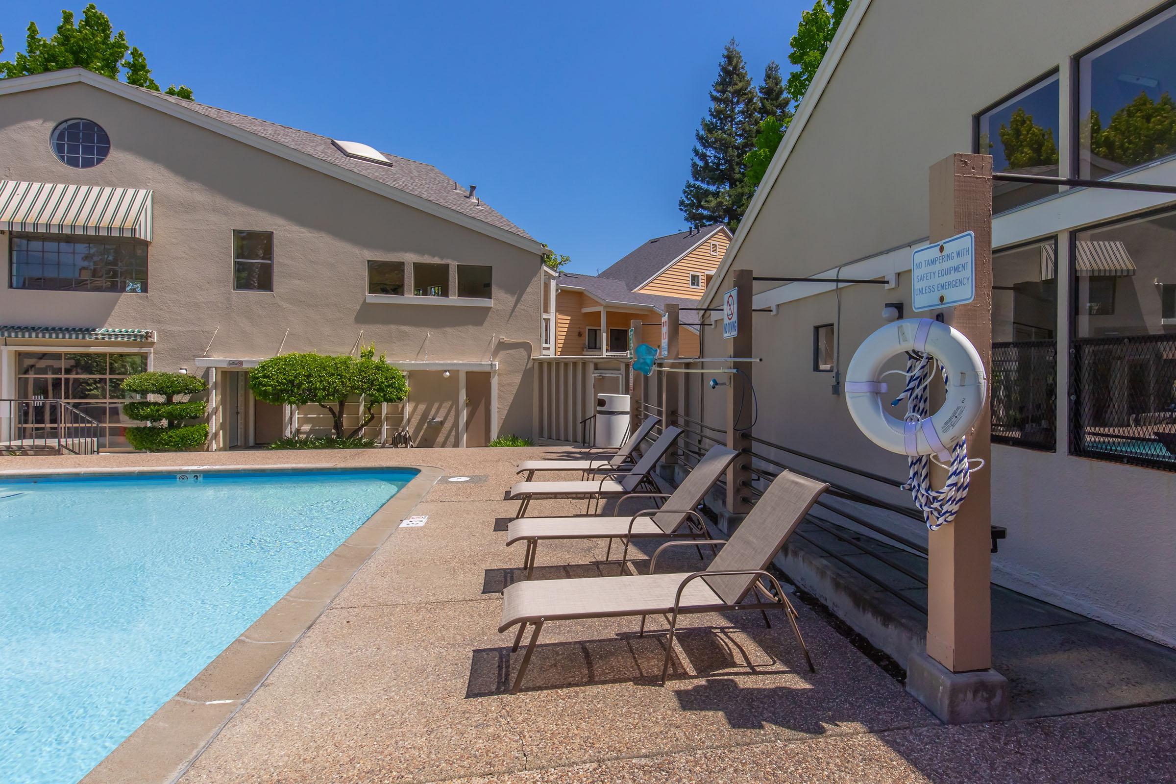 A sunny pool area featuring a clear blue swimming pool, lined with tan lounge chairs. Surrounding the pool are two buildings with light-colored exteriors and trees in the background. A life preserver hangs on a post nearby, indicating safety measures. The scene conveys a relaxing atmosphere.