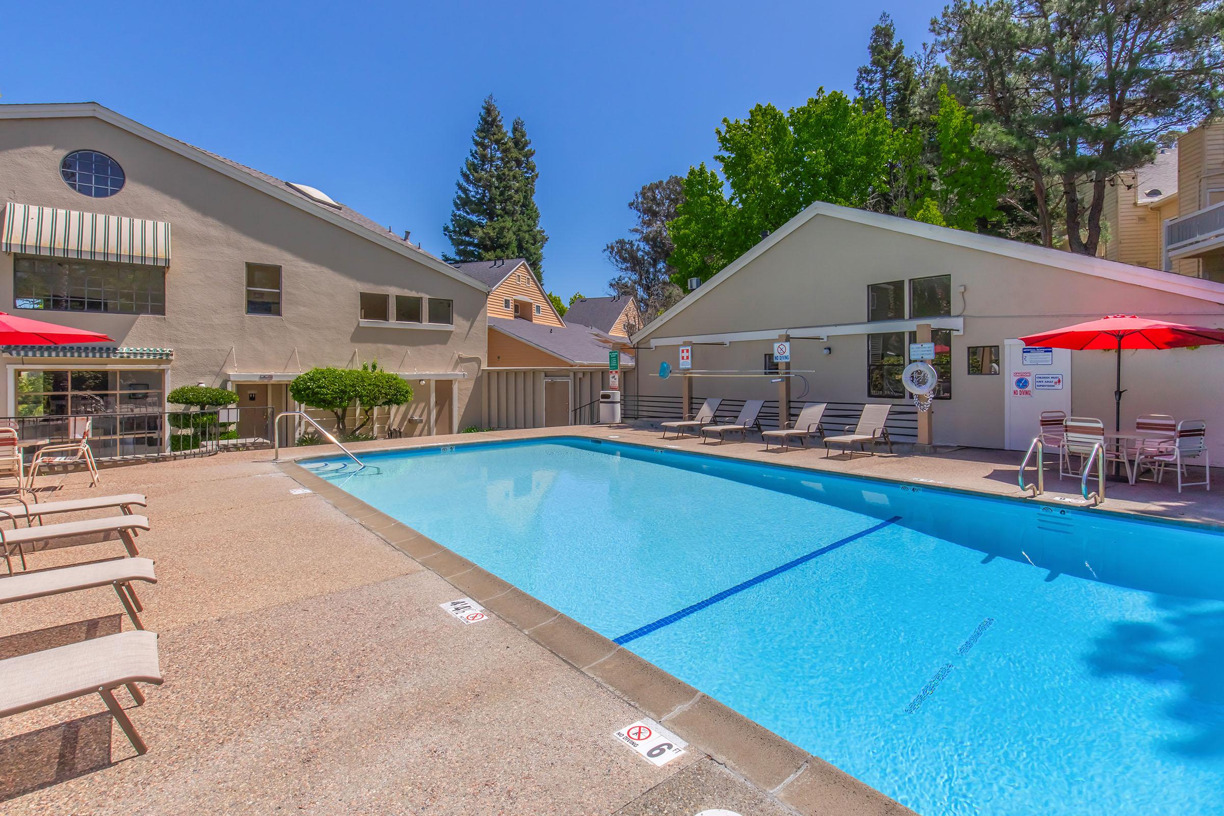 A clear blue swimming pool surrounded by lounge chairs and red umbrellas, with two buildings in the background. The area is sunny, featuring trees nearby and a calm, inviting atmosphere suitable for relaxation.