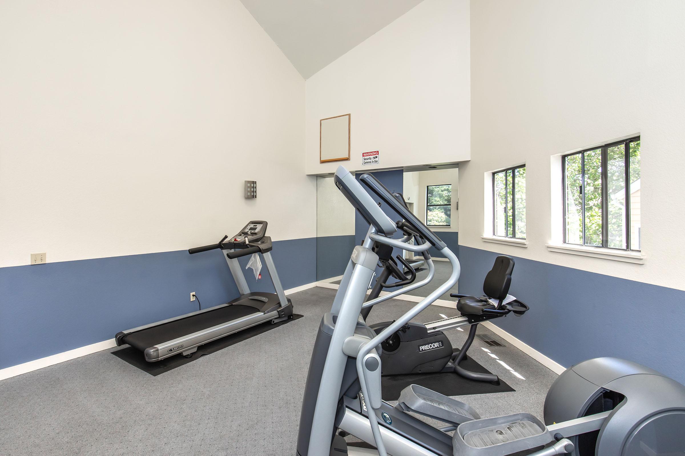 A well-lit indoor gym with a high ceiling. The room features a treadmill, an elliptical machine, and a recumbent bike, all arranged on a gray carpet. The walls are painted white and blue, with windows allowing natural light. There is a small wall-mounted control panel.