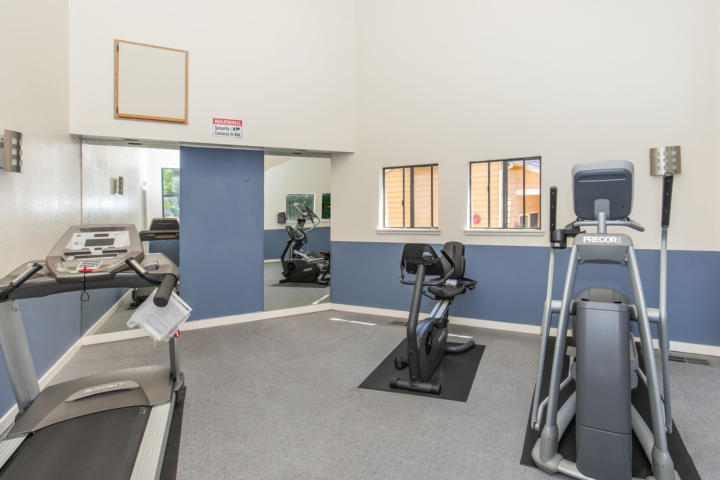 A small gym featuring a treadmill and an elliptical machine. The room has a light interior with blue accent walls, a large mirror, and windows allowing natural light. A warning sign is mounted on the wall, and fitness equipment is neatly arranged on the carpeted floor.
