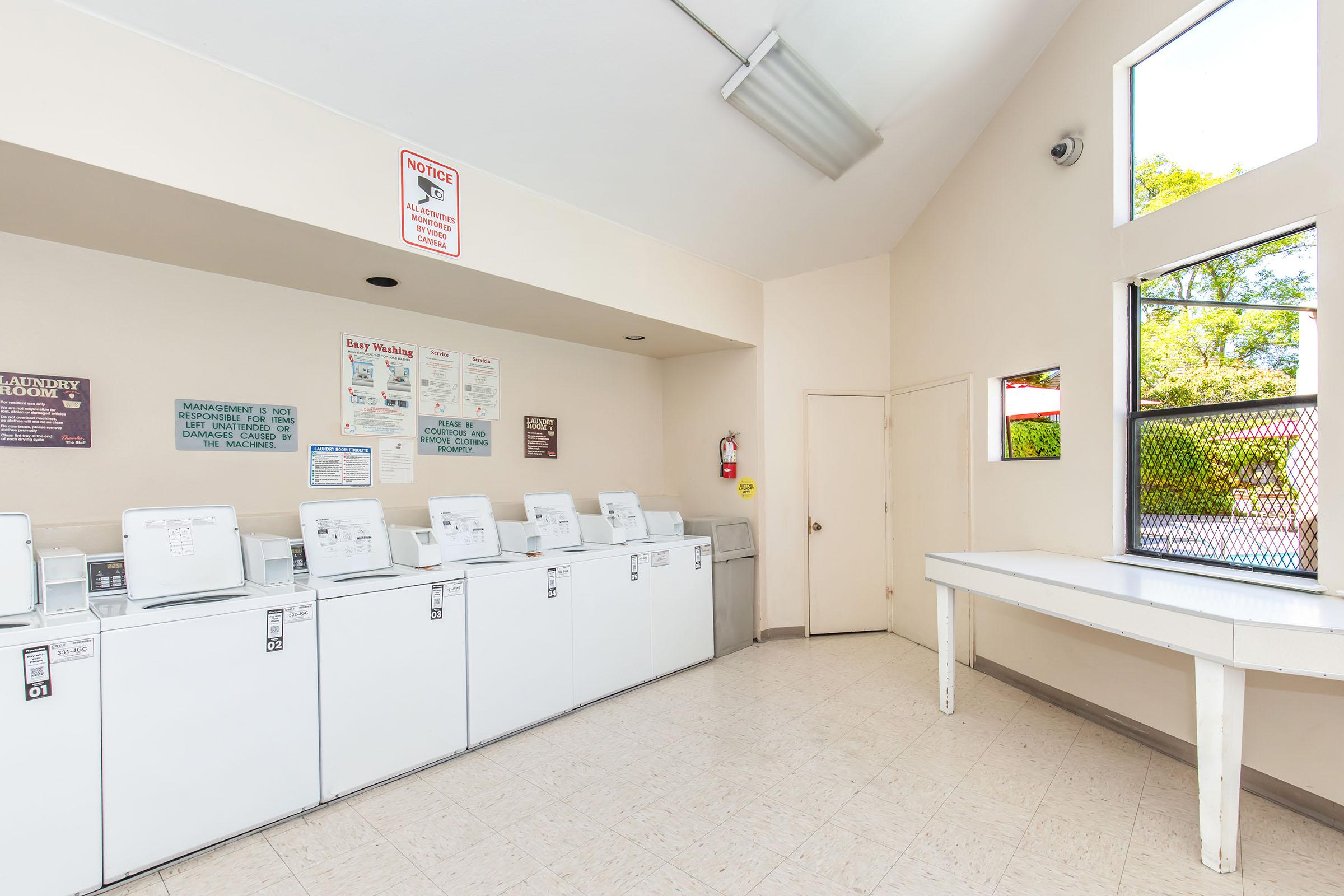 A clean and well-lit laundromat featuring several white washing machines lined against the wall. Posters with laundry instructions are displayed on the wall, and a folding table is located near the window. The space has a neutral color scheme and large windows that let in natural light.