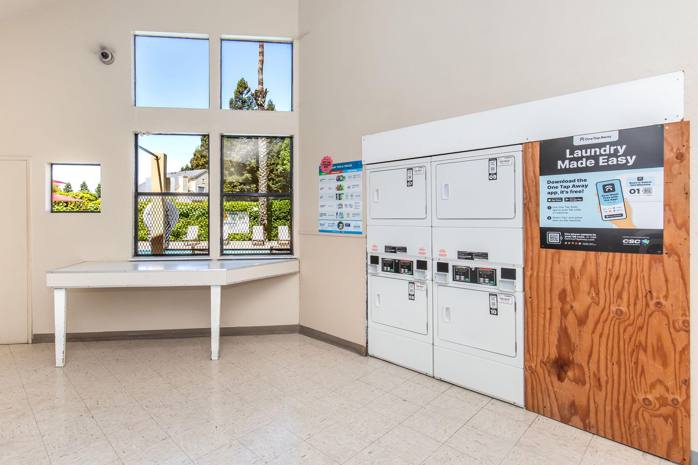 A clean laundry room featuring several coin-operated washing machines and dryers on a wooden panel, a table for folding clothes, and large windows allowing natural light in. There are informational posters on the walls regarding laundry services, and the overall ambiance is bright and tidy.