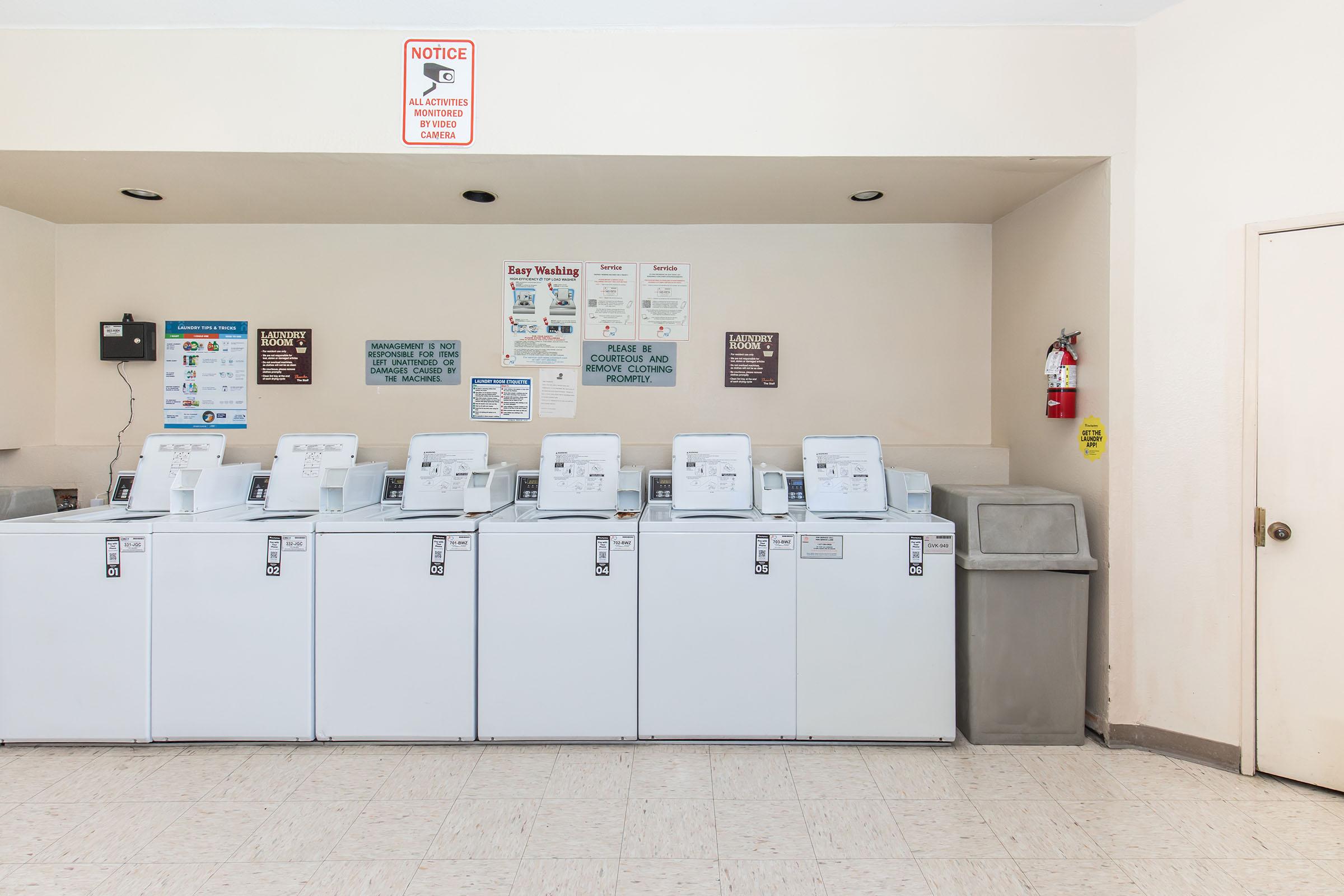 A clean laundry room featuring multiple white washing machines arranged in a row. There are notices on the wall with instructions and warnings, a fire extinguisher visible in the corner, and a trash can nearby. The floor is tiled in a light color.