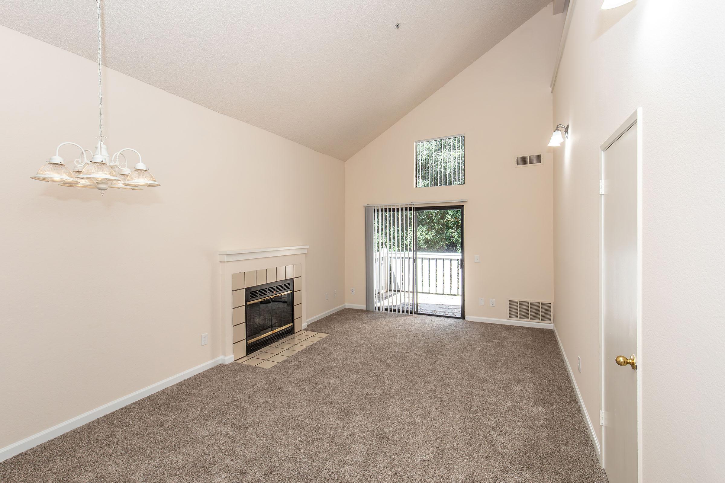 A spacious living room featuring beige walls, a high vaulted ceiling, and large windows providing natural light. There's a cozy fireplace with a white mantel, and the floor is covered in plush beige carpet. A sliding glass door leads to an outdoor space, enhancing the room's openness.