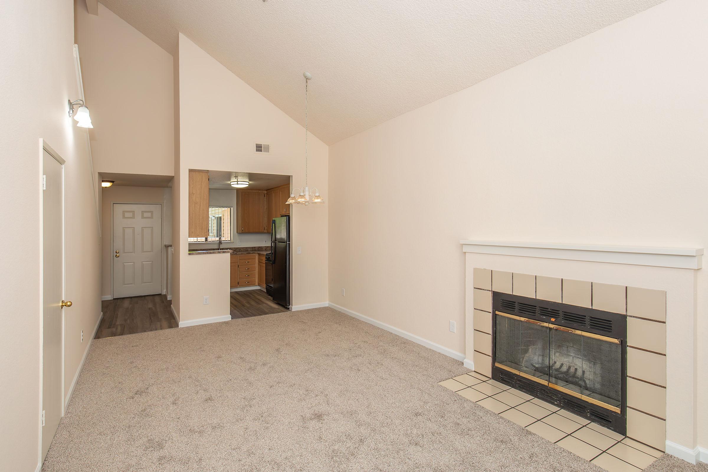 Spacious living area with beige walls and carpet, featuring a fireplace with a beige tiled surround. To the left, a doorway leads to an entryway, while in the background, a kitchen is visible. A chandelier hangs from the ceiling, and large windows allow natural light to fill the room.