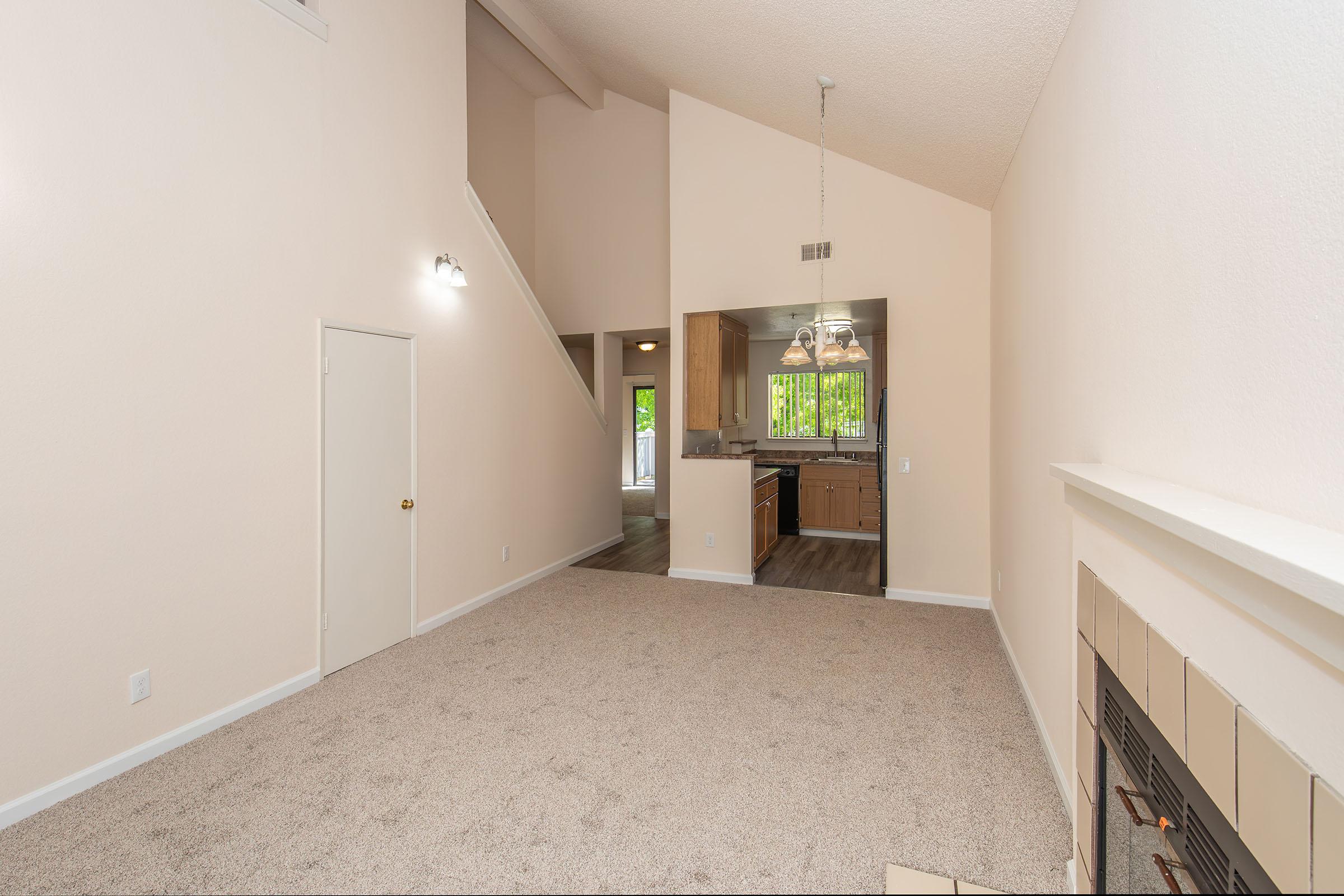 Spacious living area with neutral-colored walls and carpet, featuring a fireplace and a large window. Stairs lead to an upper level, and a doorway connects to a kitchen area with wooden cabinets and a dining area. Natural light enhances the inviting feel of the space.
