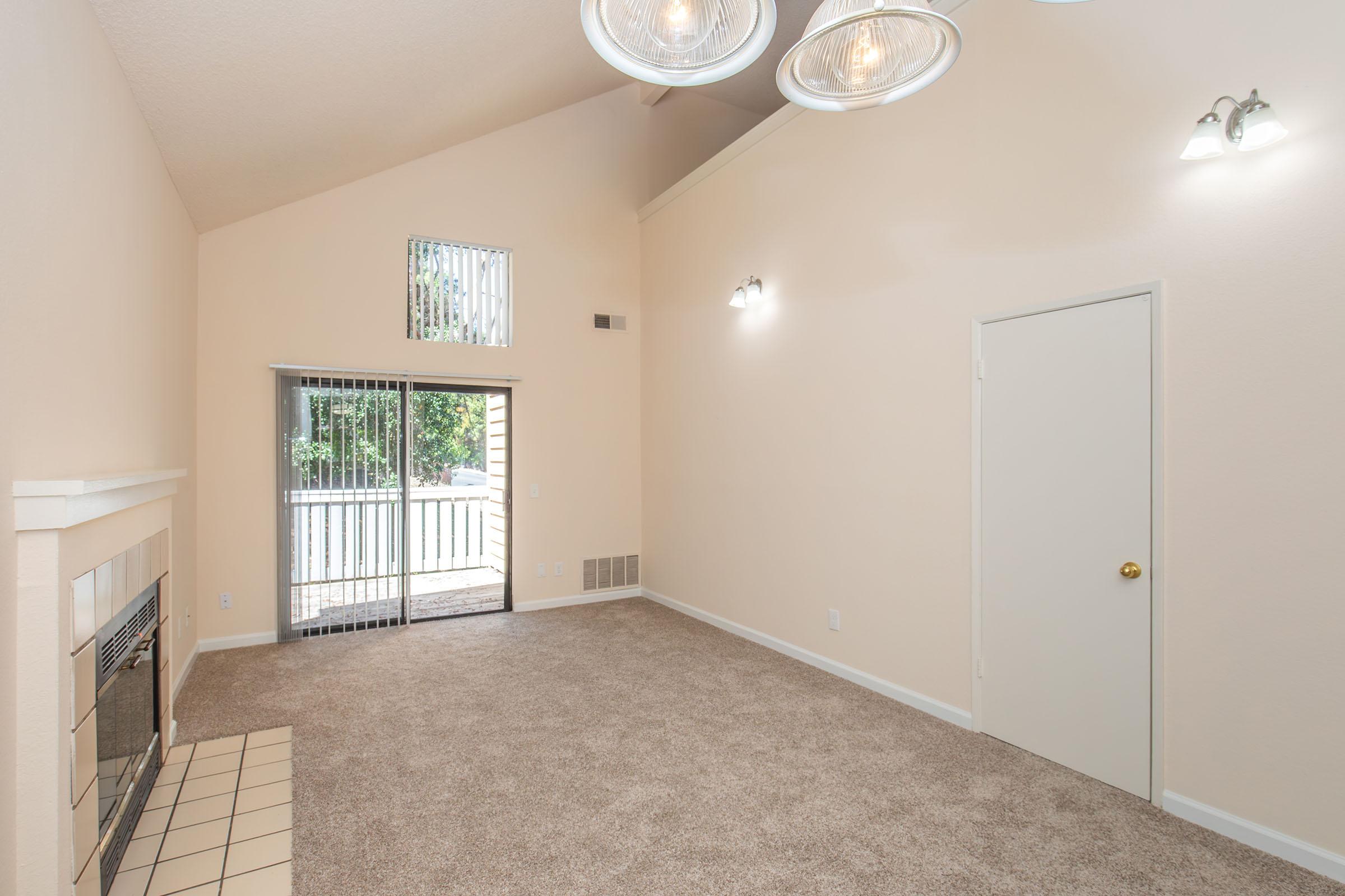 A spacious living room featuring light beige walls and carpet, with a large window allowing natural light from outside. There is a fireplace on one wall and modern light fixtures hanging from the ceiling. A door leads to an outdoor patio area, creating an inviting atmosphere.