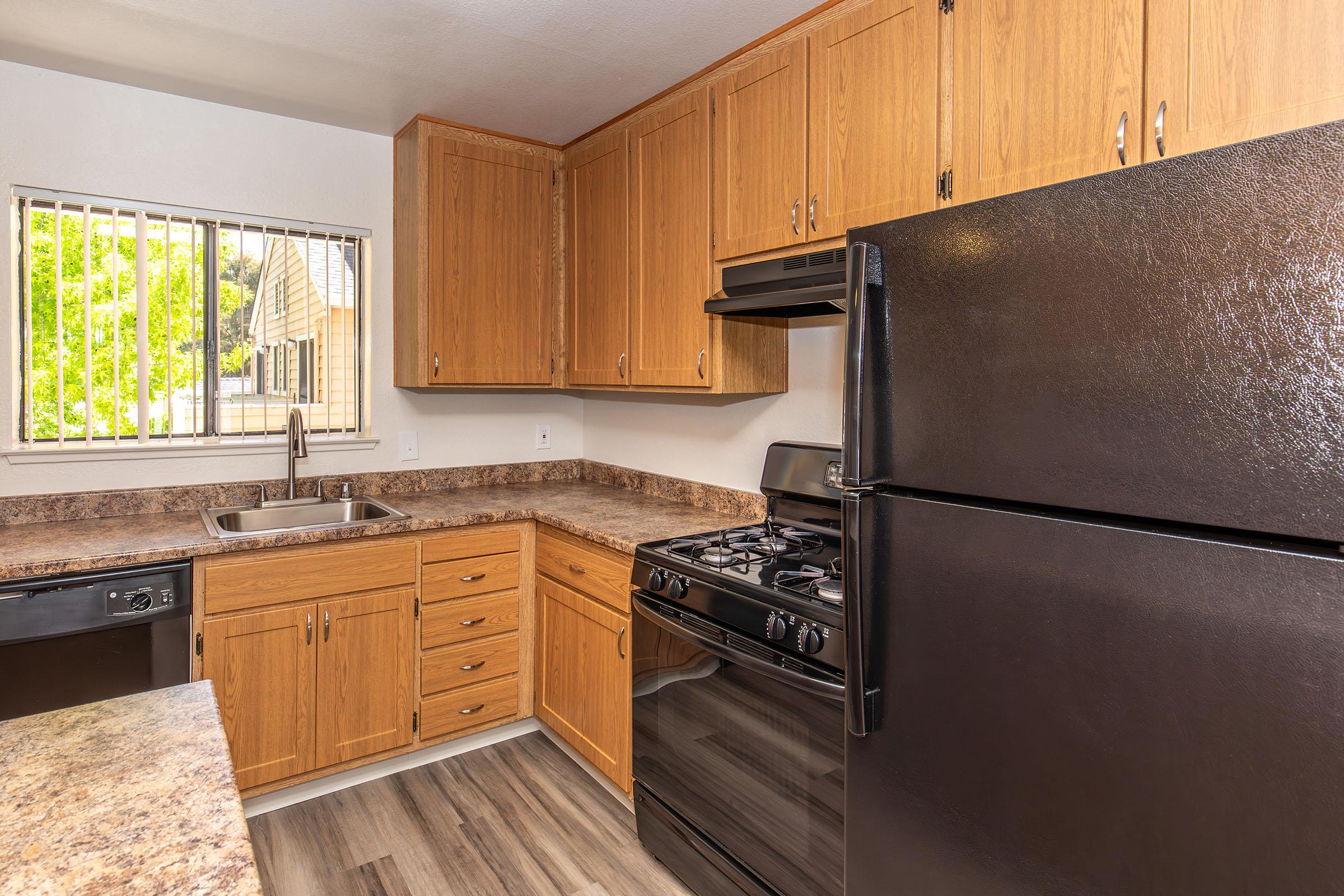 A modern kitchen featuring wooden cabinets, a black refrigerator, and a black gas stove. The countertop is a light-colored stone, and there is a stainless steel sink beneath a window. The flooring is wood-like laminate. Natural light comes in through the window, illuminating the space.