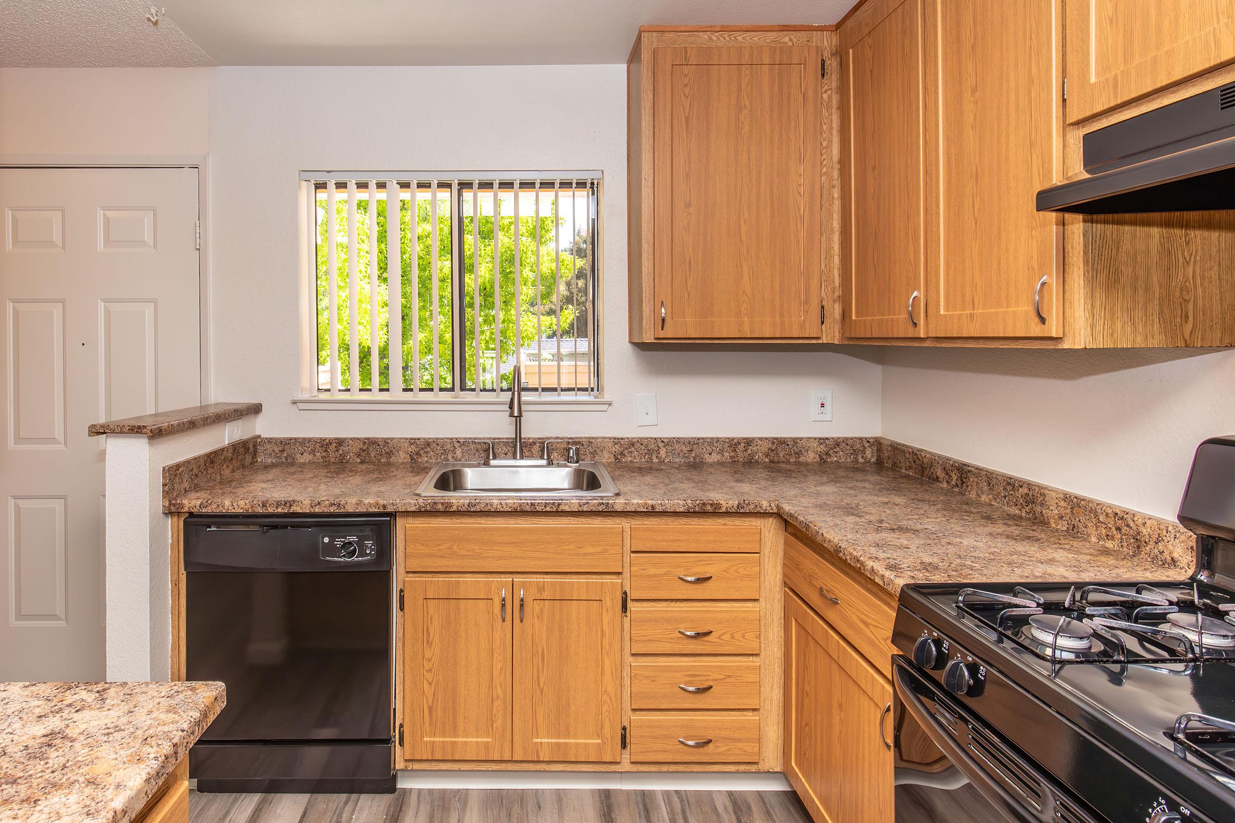 A bright kitchen featuring wooden cabinets, a stainless steel sink, a black dishwasher, and a gas stove. The countertop is made of a speckled brown material, and there is a window above the sink allowing natural light to enter. The flooring is a modern gray laminate.