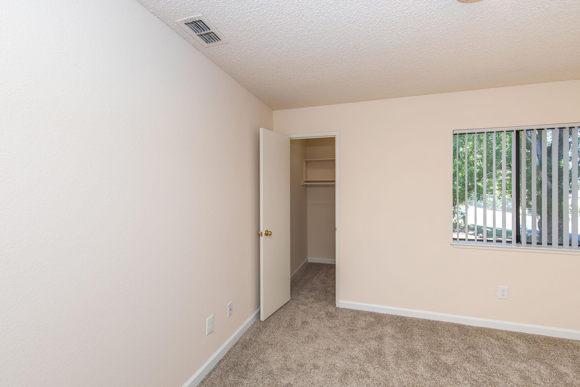 A view of an empty room featuring light-colored walls, a beige carpet, a closet with an open door, and a window with vertical blinds allowing natural light. The room has a simple design, making it suitable for various uses or as a bedroom.