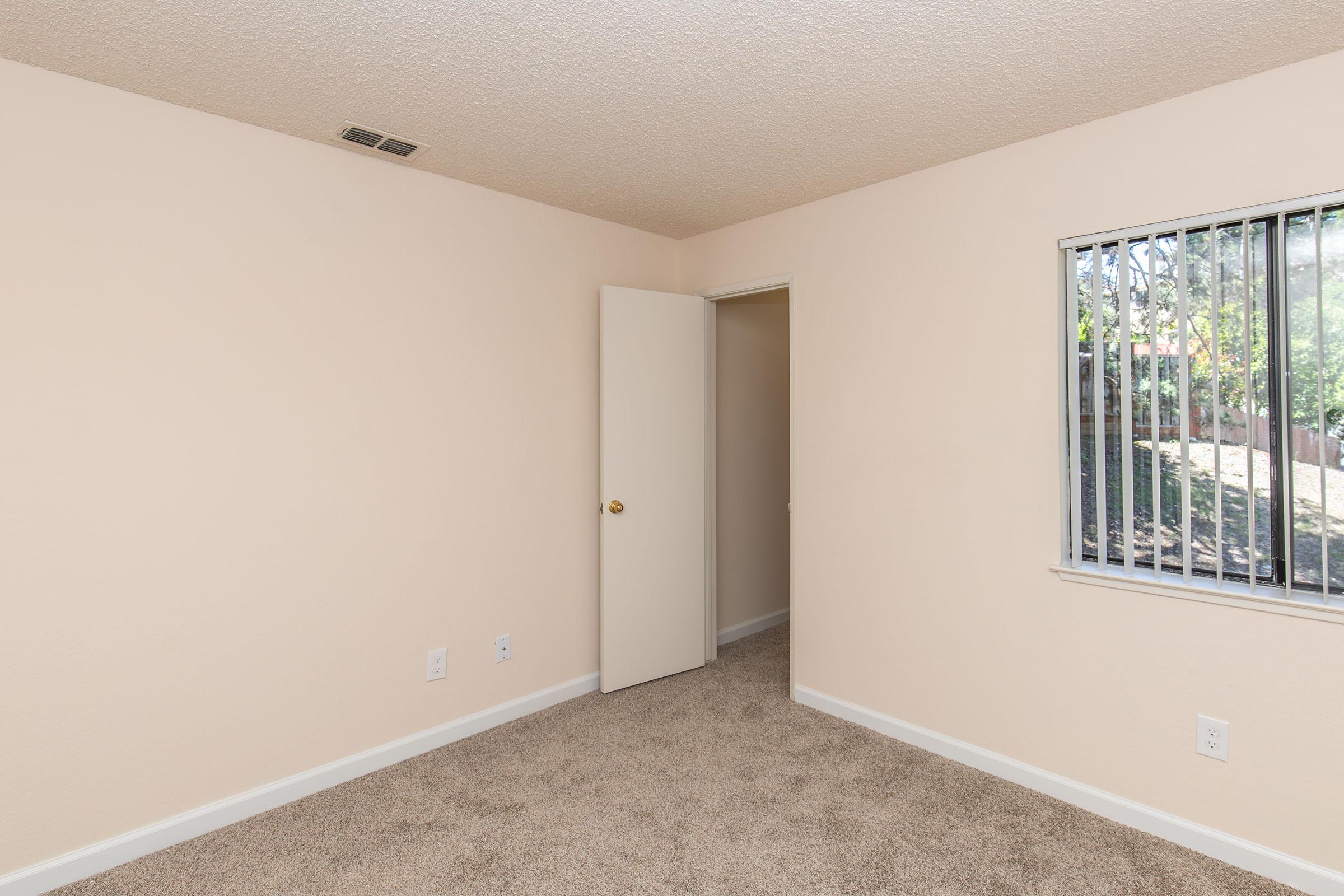 A vacant room with light beige walls and carpeted floor. There is an open door leading to another area and a window with blinds allowing natural light to enter. The space appears clean and simple, ideal for use as a bedroom or office.