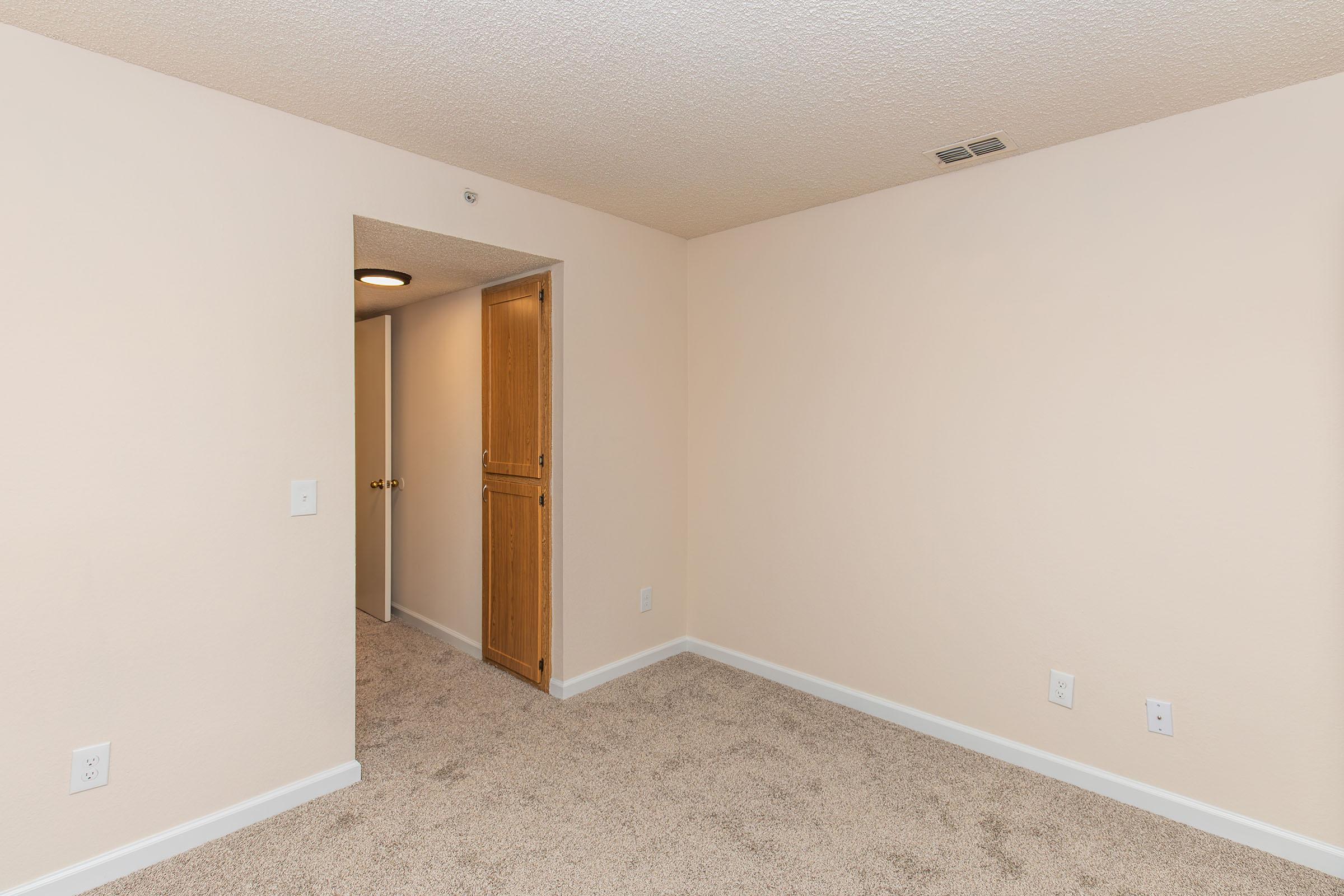 A clean, empty room with beige walls and a textured ceiling. The floor is covered with light-colored carpet. There is an open doorway leading to a hallway, and a closet with wooden doors is visible on the left side. The atmosphere is bright and spacious, suggesting potential for various uses.